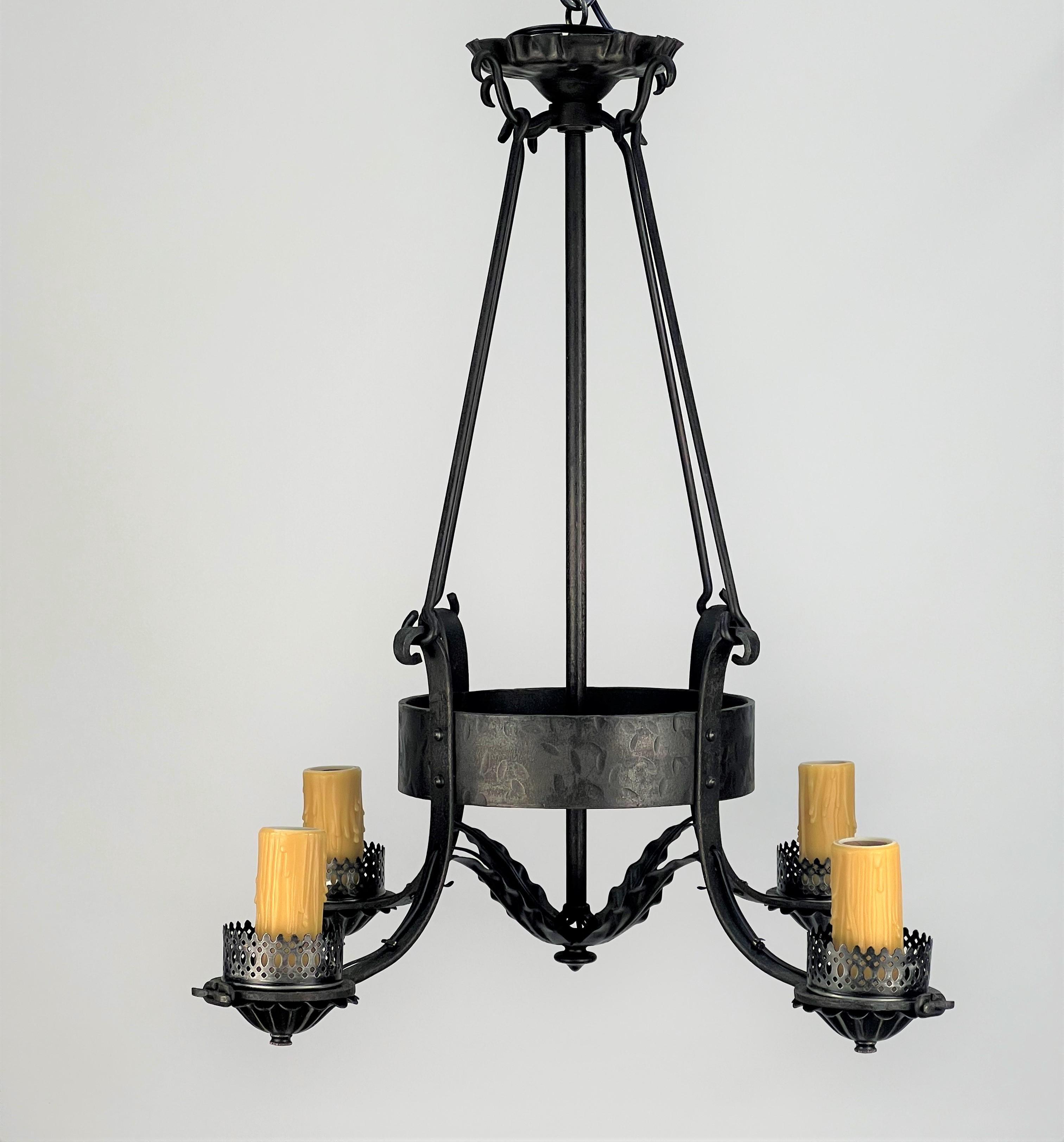 A rustic 1920's American wrought iron chandelier featuring four candle base sockets with faux beeswax drip candle sleeves. Each of the candles are attached to an arm featuring a reticulated (pierced) gallery on top and a shell shaped cup underneath