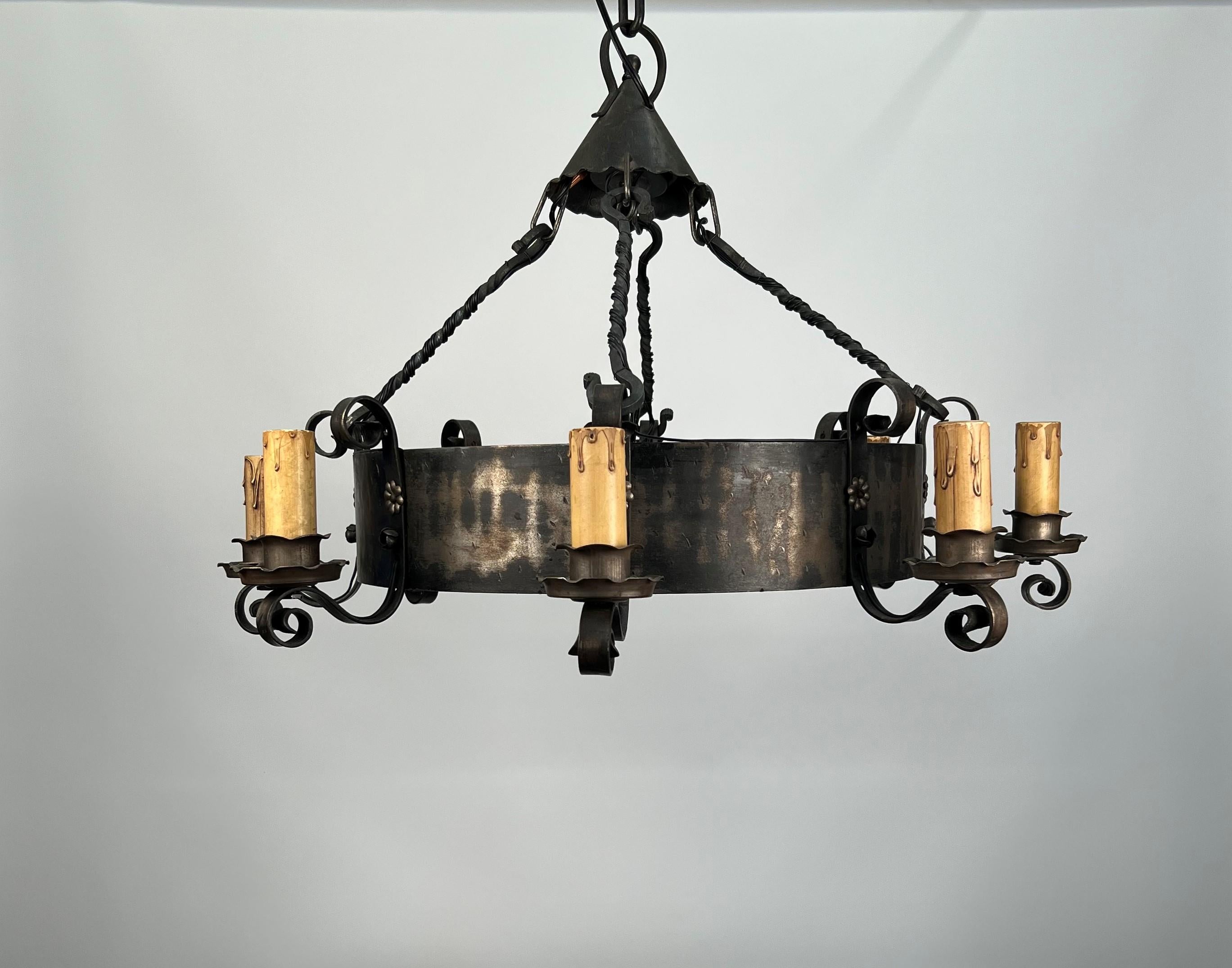 A rustic 1920's American wrought iron chandelier featuring eight medium base candle sockets with original drip candle sleeves. Each of the candles are attached to a central steel strap by a stamped steel rosette and rose holding decorative iron