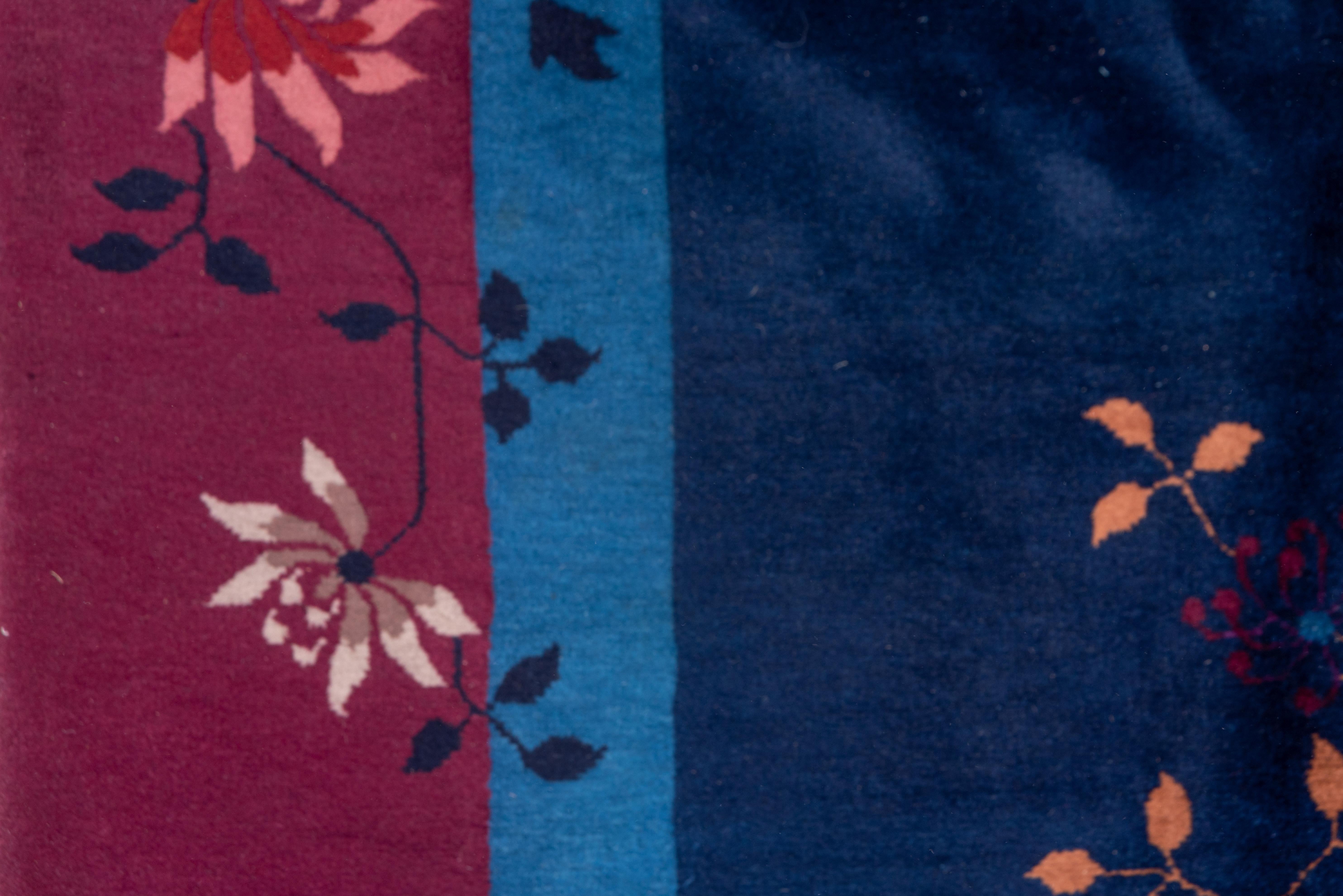Ce tapis urbain de l'entre-deux-guerres à tissage moyen présente un champ bleu royal centré sur un motif géant polychrome de fleurs et de tiges, avec des oiseaux et des papillons qui voltigent. Vase et pots dans la bordure profonde de framboise. Des