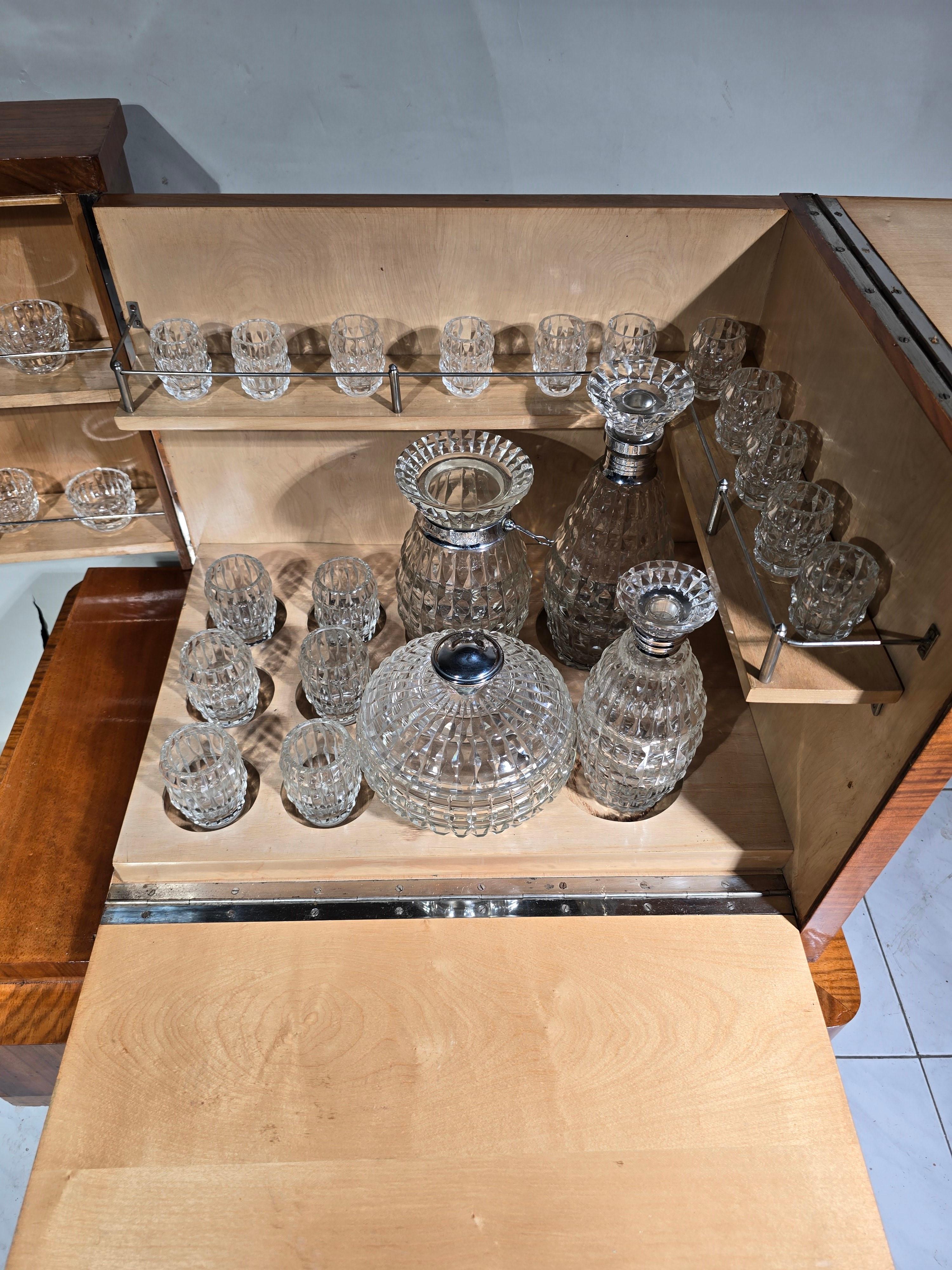 Add a touch of timeless elegance to your home with this stylish Art Deco bar cabinet from the 1920s. Crafted from exotic wood, this exquisite piece exudes sophistication and luxury.

Featuring a set of cut glassware including various containers and