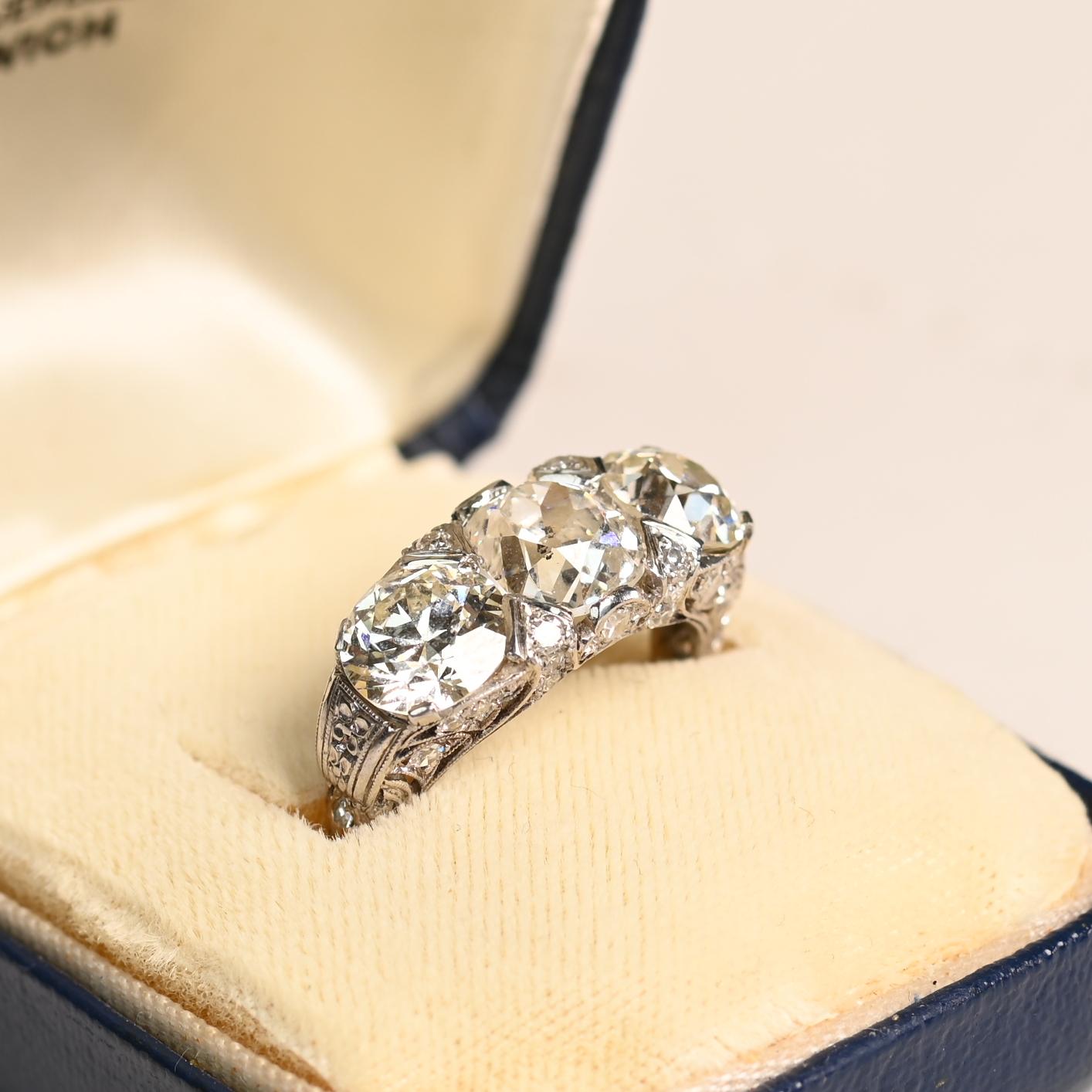 Este exquisito anillo Art Déco de tres piedras es un verdadero testimonio de la artesanía de principios del siglo XX. En su corazón yace un impresionante diamante antiguo talla cojín, de 2,22 quilates, con un cálido color J y claridad SI2.