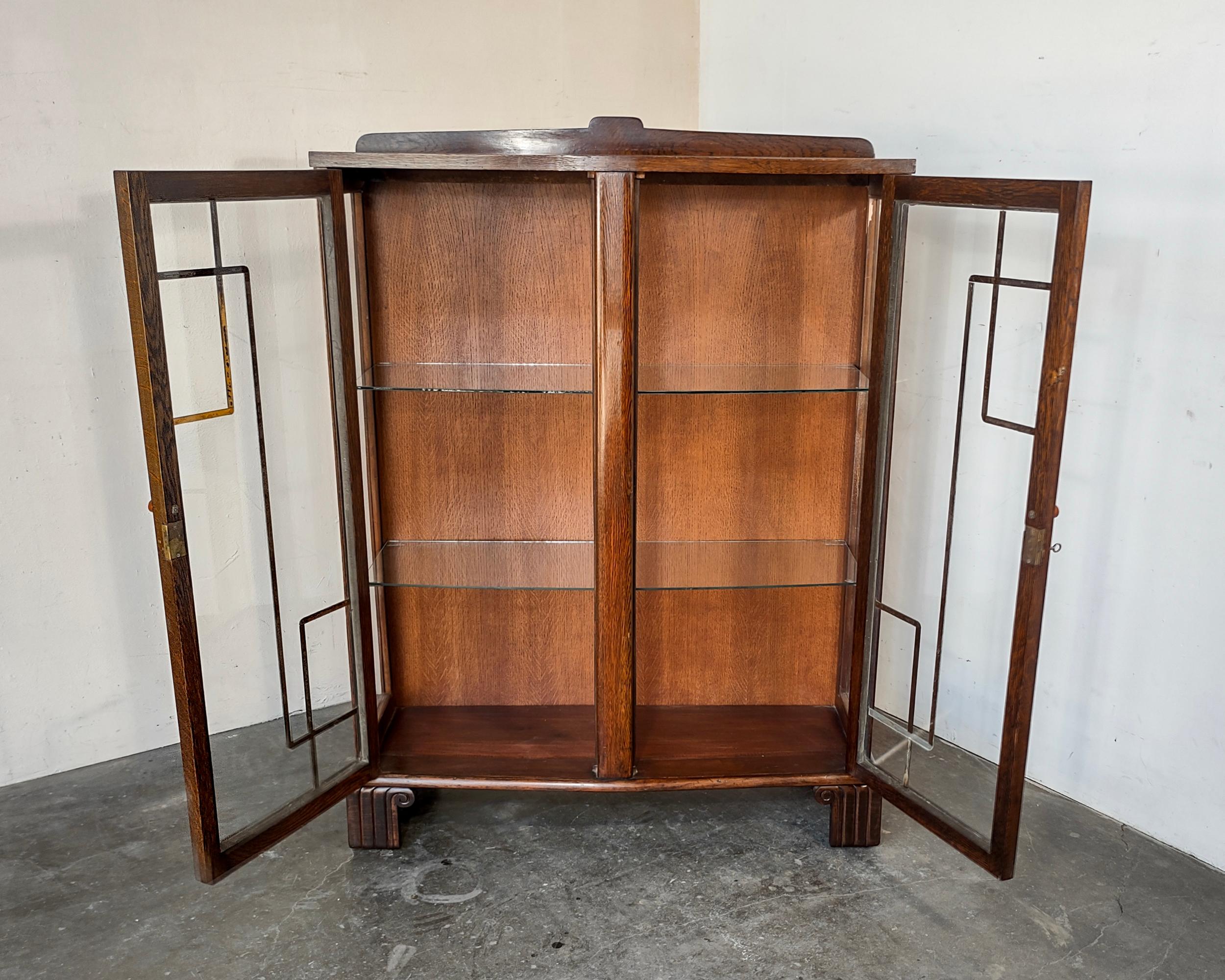 1920s Art Deco glass display cabinet featuring two slightly curved glass shelves inside. Beautiful design details include column feet, mirrored glass design on doors and bakelite pulls. Original locking key included. Overall great vintage condition