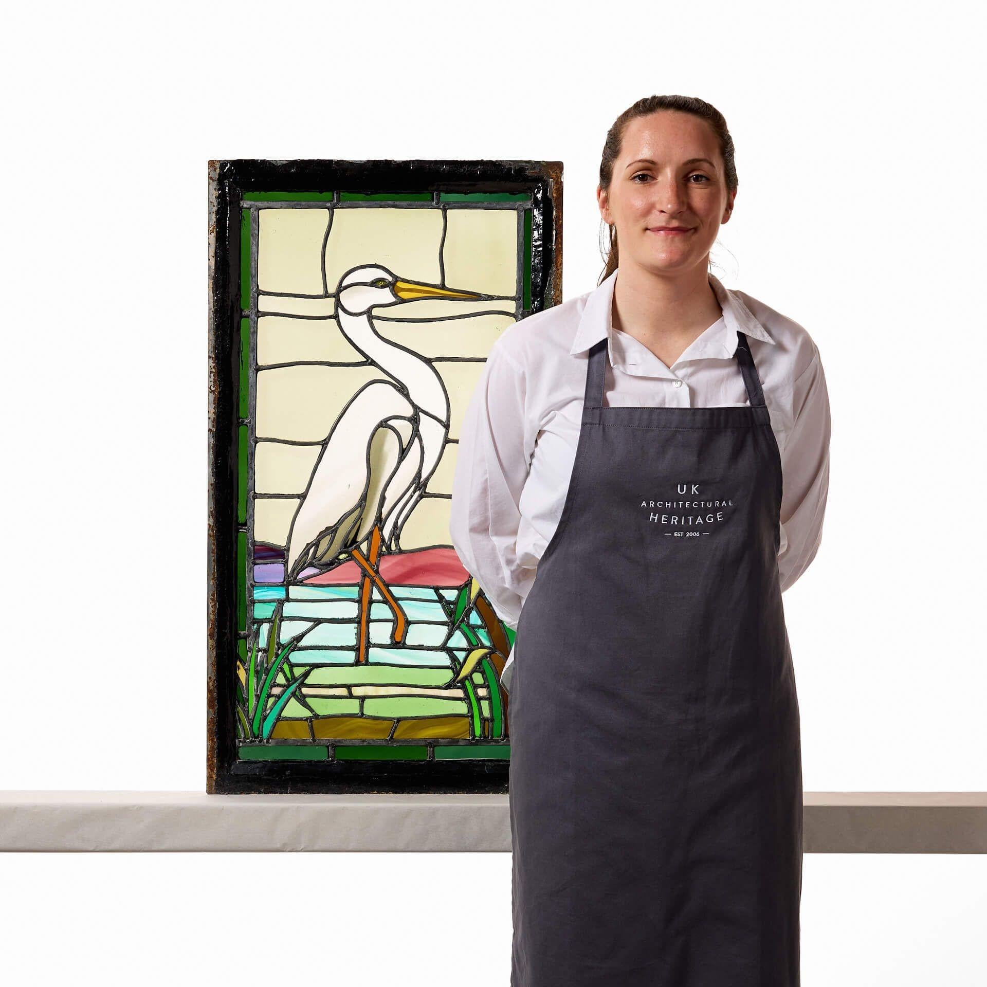 This idyllic 1920s Art Deco stained glass window depicts a great white heron (also known as a great egret) standing gracefully on the river bed.

Tall green reeds grow on the riverbank, while sloping panes of stained glass beyond the blue water of