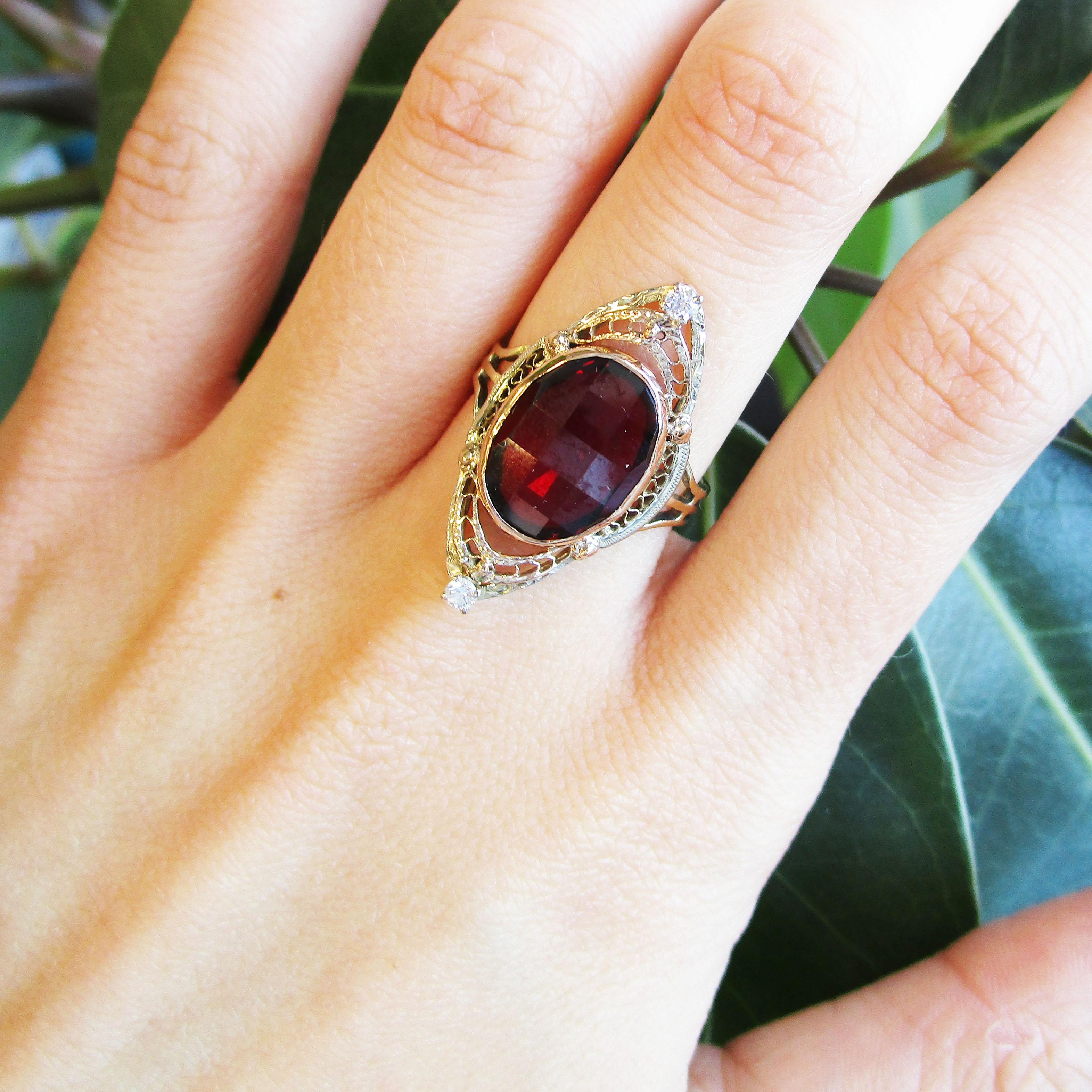 This is an enchanting Art Deco ring featuring two colors of 14k gold in a dramatic, long layout with a garnet center and two delicate diamond accents. The garnet has a finely detailed filigree border in rose and white gold that come together to form