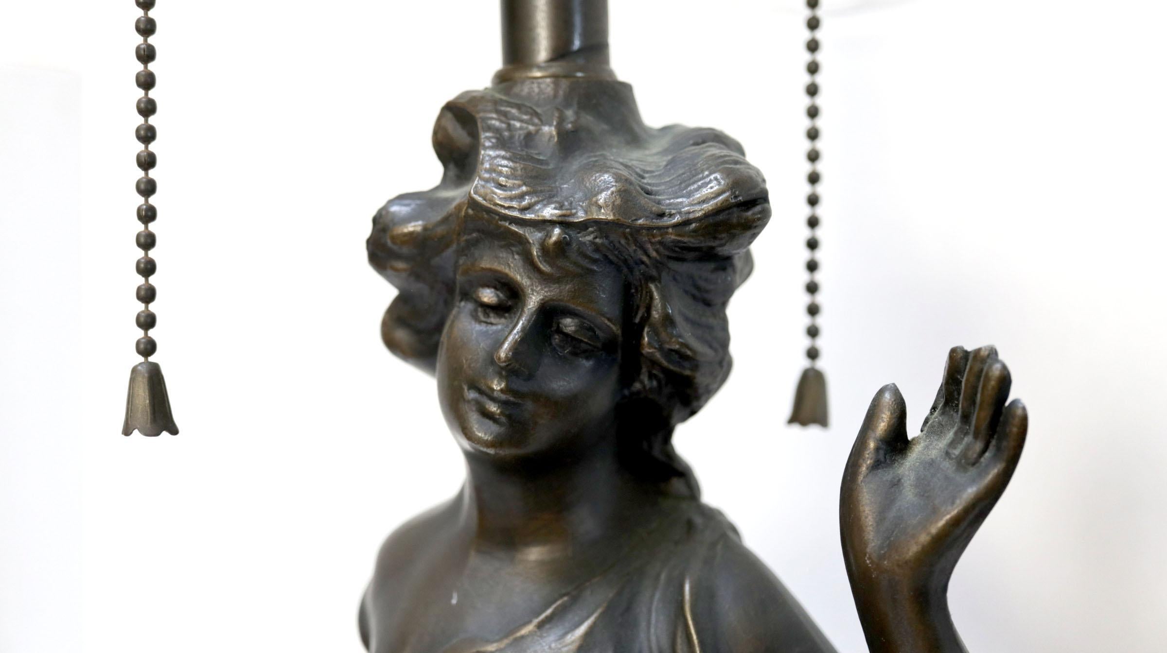 An antique Art Nouveau style bronze figural table lamp is from Europe circa 1925. It is in the French taste in the form of a woman in classical drapery standing on one foot on a tall circular section base with floral detailing. The lamp has two