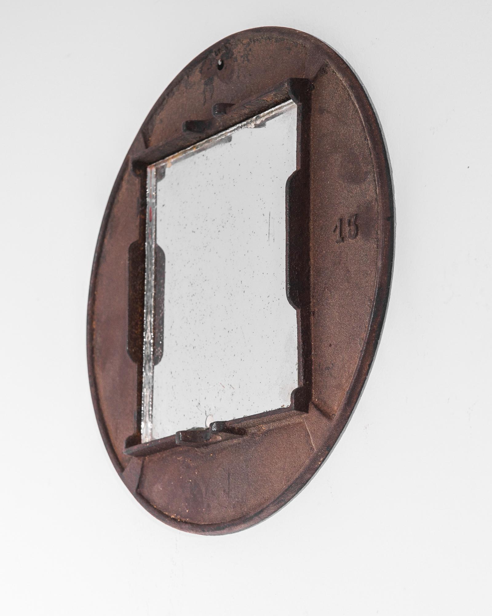 An iron mirror from 1920s Belgium. An interplay of shapes and textures creates a weathered industrial aesthetic: a rectangular mirror is set inside a circular iron frame, oxidized to a dusky burgundy. The soft patina of the metal and the chipped