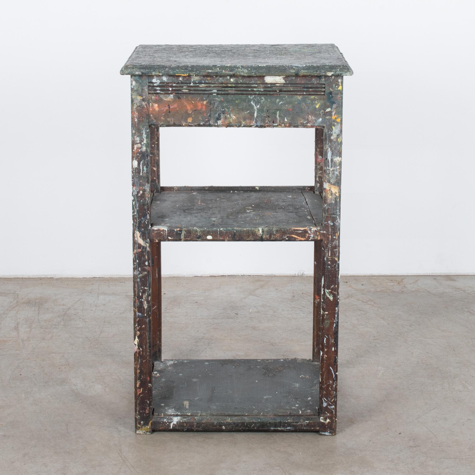 A painters table from Belgium, circa 1920. A square painter’s box, originally used for storing paints and brushes, sits upon wooden legs. The lid of the painter’s box opens on its hinges, sustained with a slender chain. Two lower shelves provide