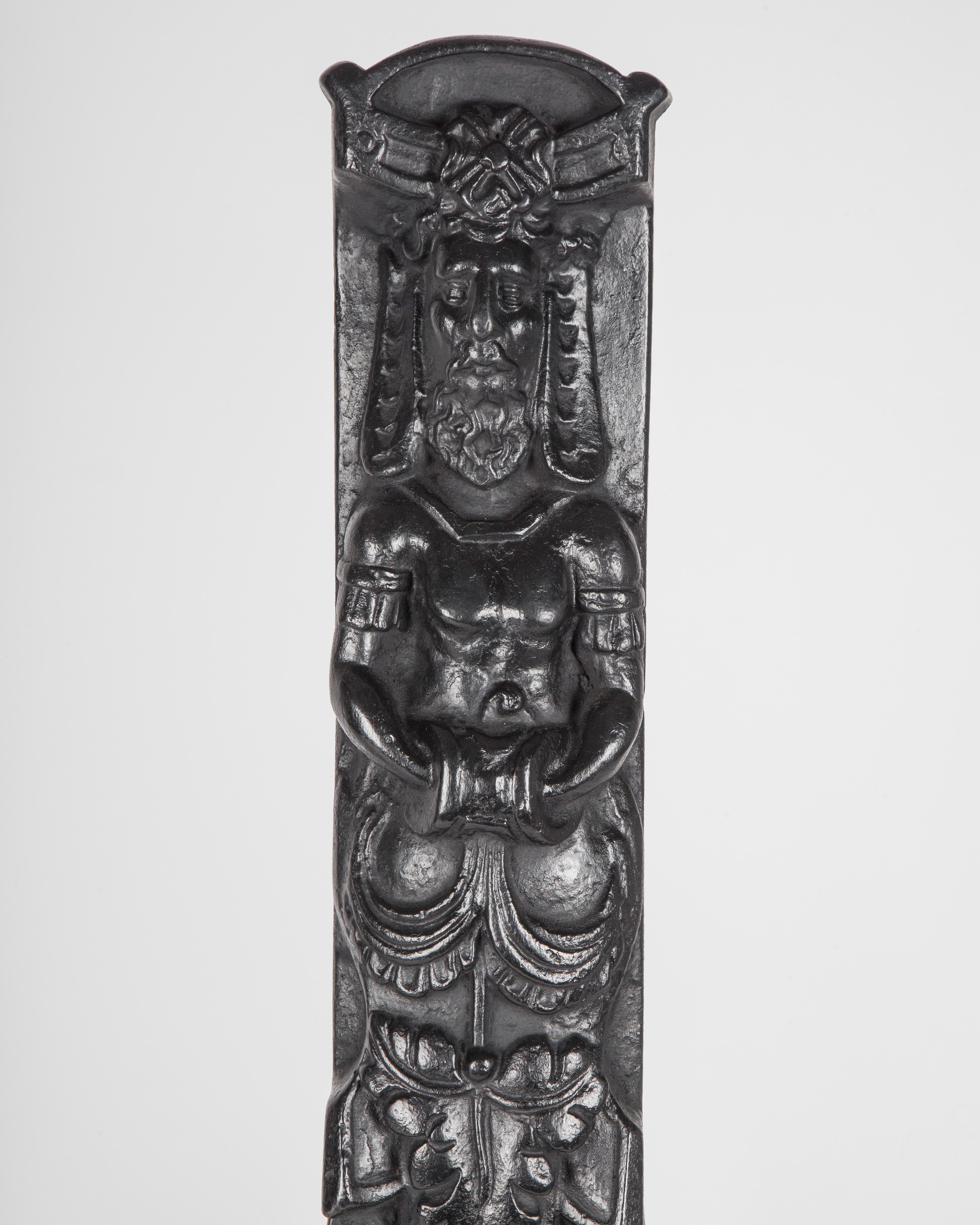 AFP0674
A pair of unusual large antique andirons each having a figure of a hybrid atlas over a caduceus in the main rectangular column, as well as masks, dolphin fish and crosses on the curves of the legs. In cast iron with a blackened finish. From