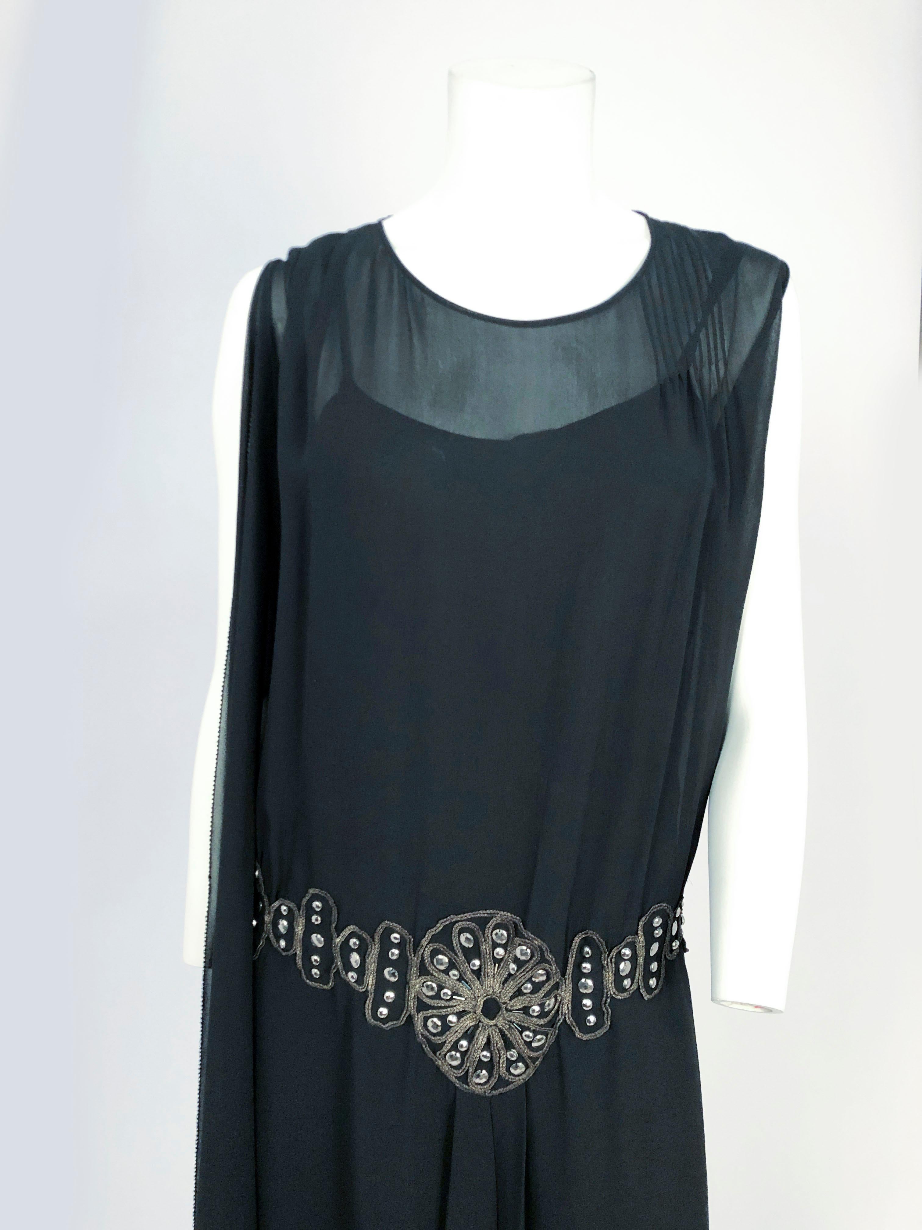 1920s Black Drop-waist Dress With Beading and Drape Accents. The hand appliqué is trimmed in lamé rope and rounded studs. The shoulder has a georgette drape and the face of the dress is pleated and open along the skirt.