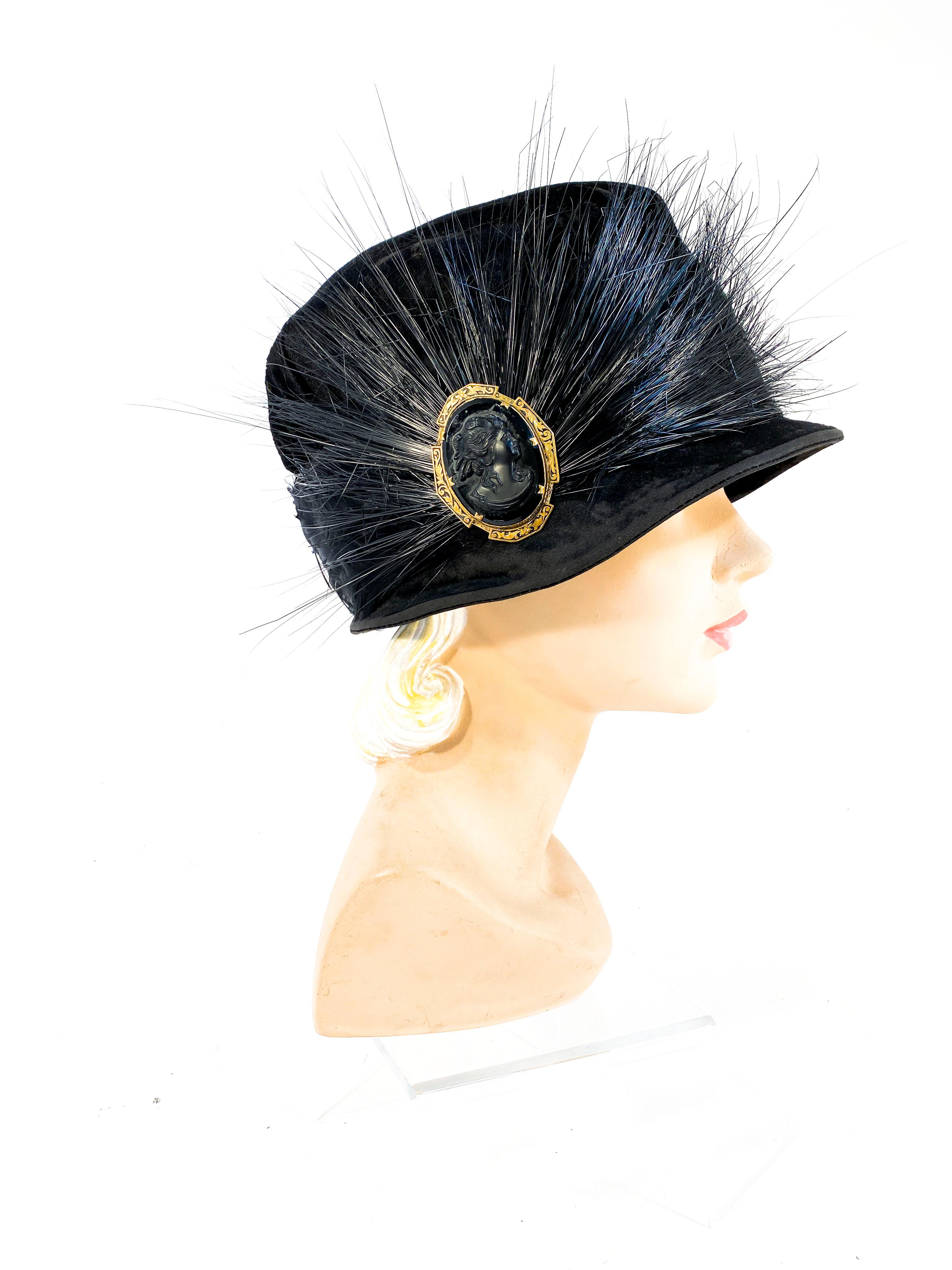 1920s black velvet cloche hat featuring a band of enlarge black bird feathers. On the side of the cloche there is a large panel of horsehair wisps centered with a black jet and brass cameo piece. The label on the interior tell us that this was a