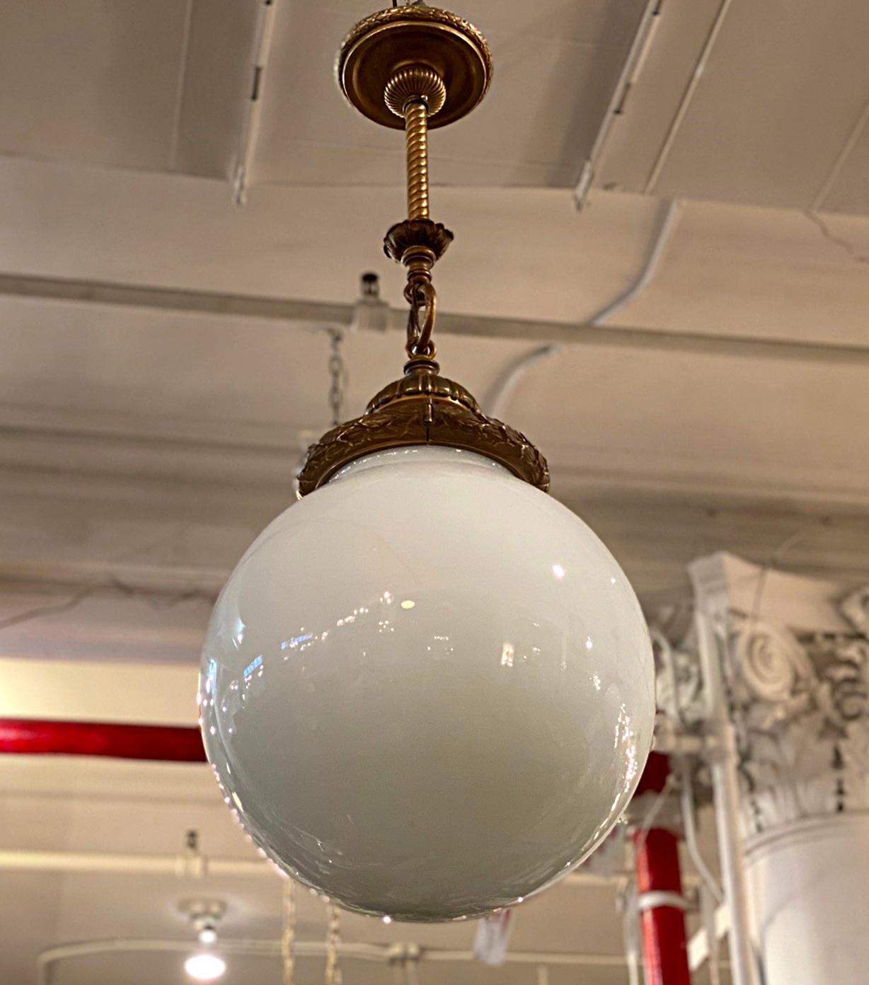 1920s brass pendant light with a large white opal glass globe. The cap has an antique brass patina with decorated wreath and ribbon design attached with a chain and tube fixture. This can be seen at our 333 West 52nd St location in the Theater