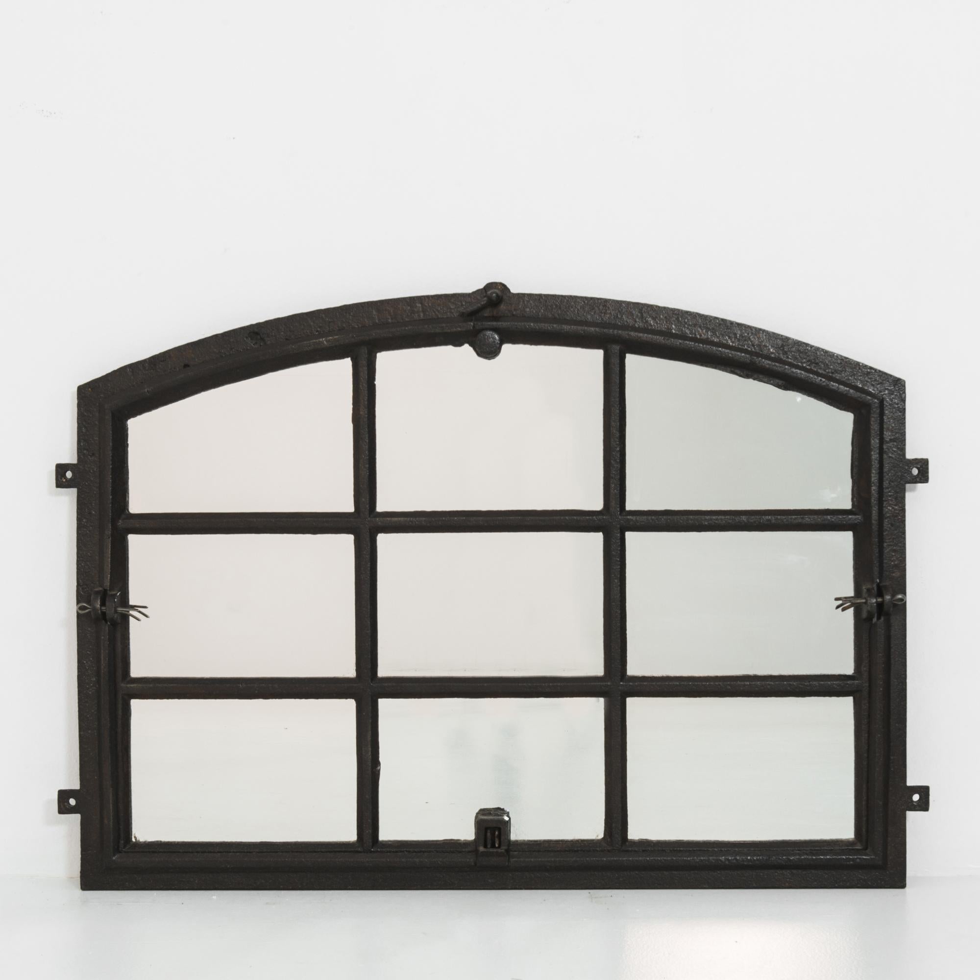 A 1920s cast iron mirror from Central Europe. Originally a factory window, the dark color and solid heft of the metal create an impressive sense of presence. The gridded panes are crowned by a wide arch; iron hinges and window fastenings provide