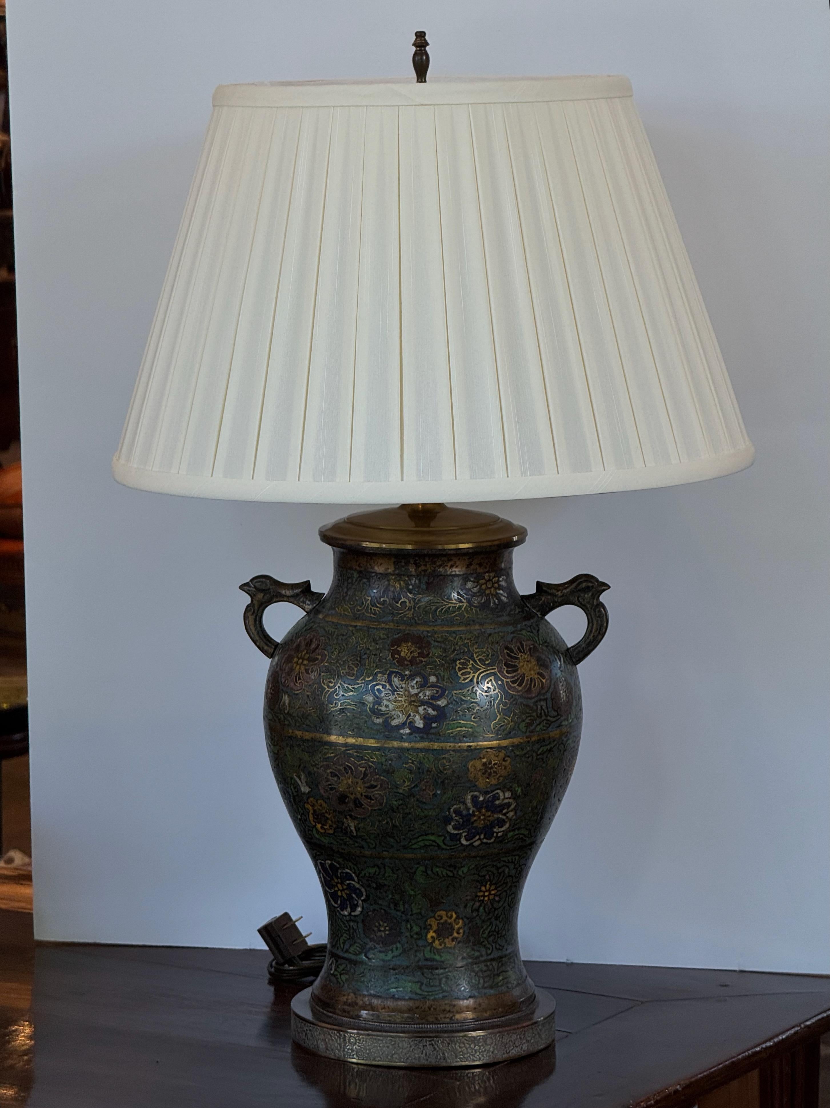 Table lamp featuring a champlevé enamel body with richly colored floral patterns in blue, green, and gold tones. The metal base and handles complement the intricate cloisonné-style detailing, topped with a pleated cream shade for a classic look.