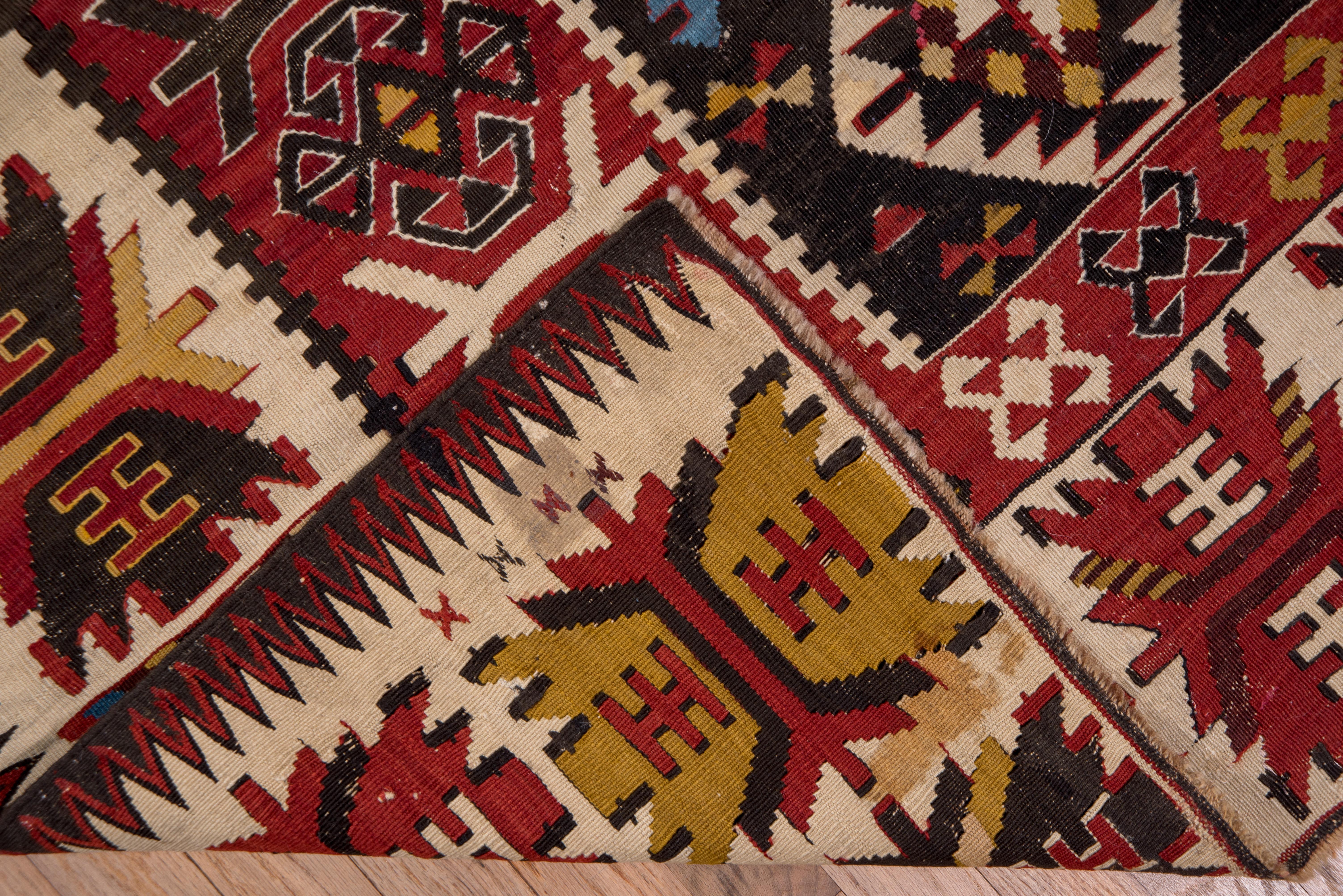 This slit tapestry piece shows a pole medallion of four hooked and nested diamonds on the near black ground, with supporting similar secondary elements, in off white, goldenrod, red, green and cerulean. Main ecru border with split, serrated