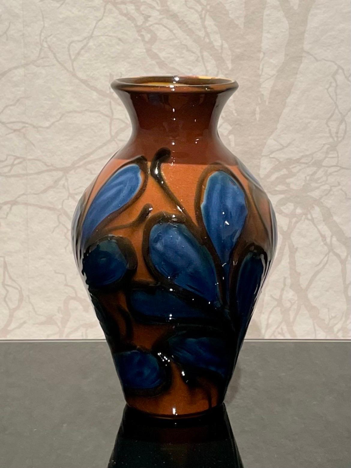 This is the 1920s Danish 22 cm high ceramic vase by Herman Kähler. 

It comes with a baluster shaped body with a glossy surface. It has a beautiful cow horn glazed pattern in musty colors. 
Black colored climbing plants with royal blue flowers on a