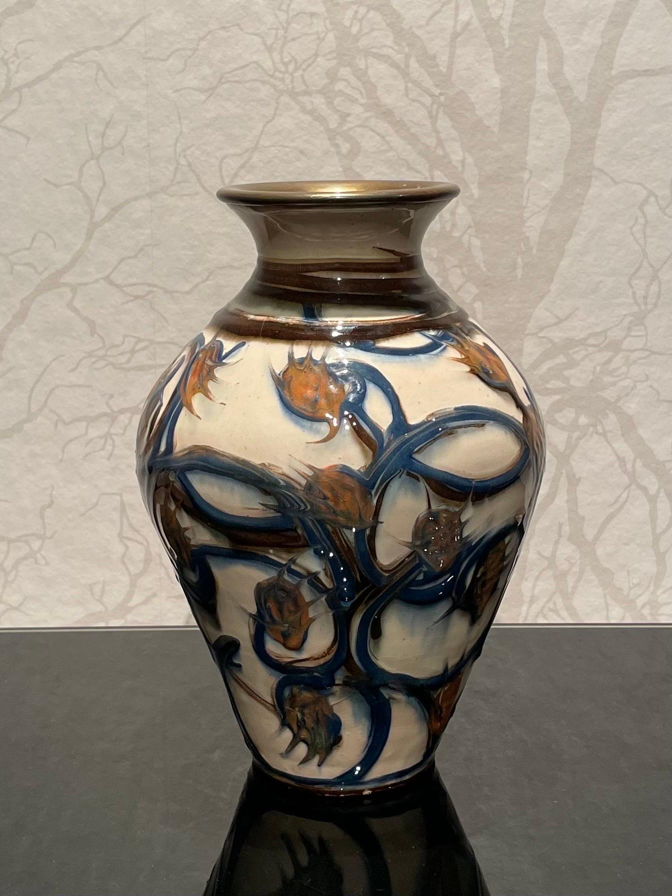 This is the 1920s Danish 23 cm high ceramic vase with a bronze colored mouth by Herman Kähler. 

It comes with a baluster shaped body. It has a glossy surface, a beautiful cow horn glazed pattern with mediterranean blue climbing plants and light