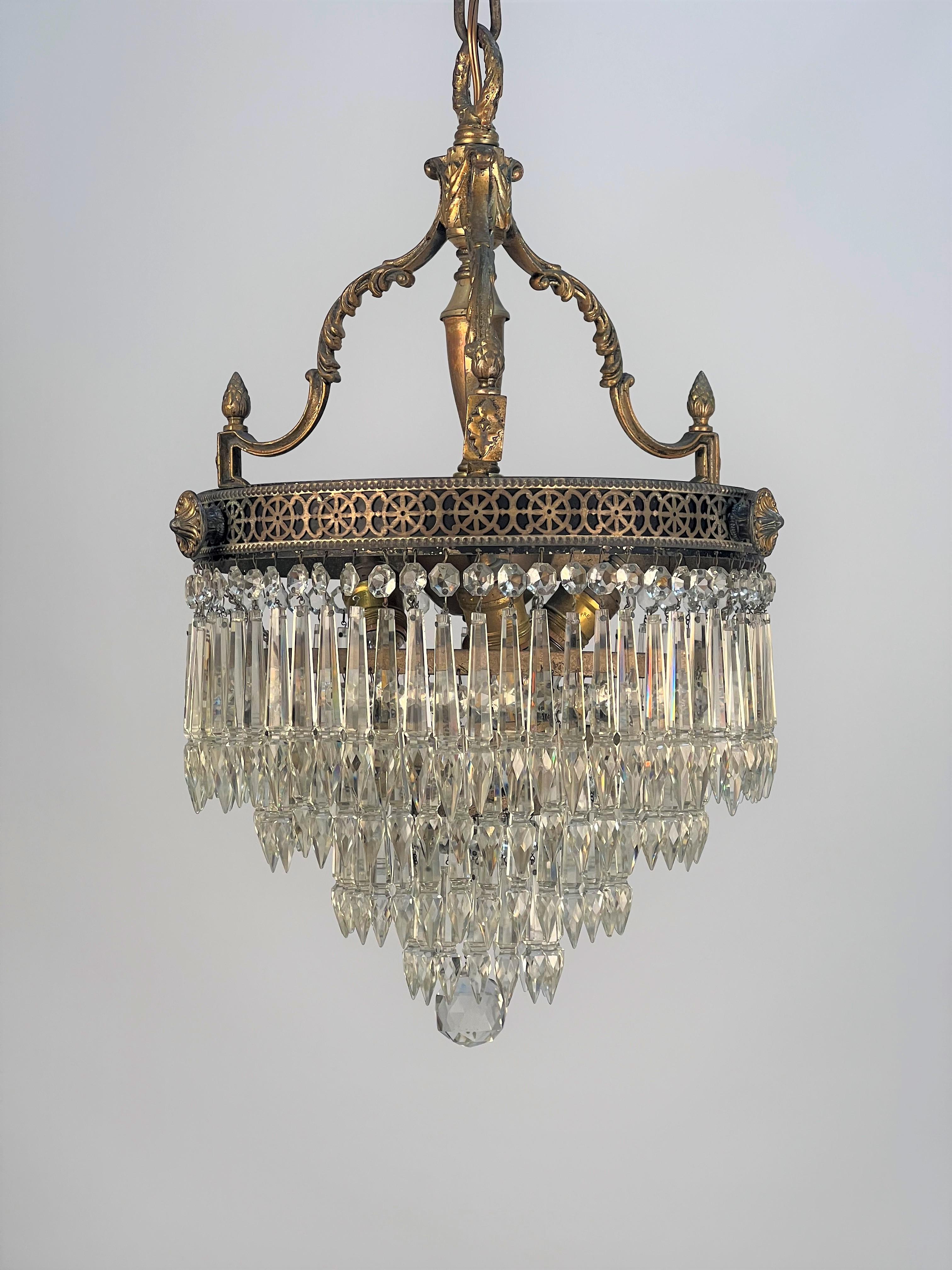 This 1920's chandelier features three cast brass decorative arms with acanthus leaf detail curving down word to a rectangular stop with a turned finial on top. The stop appears to be attached to a central reticulated (pierced) brass gallery applied
