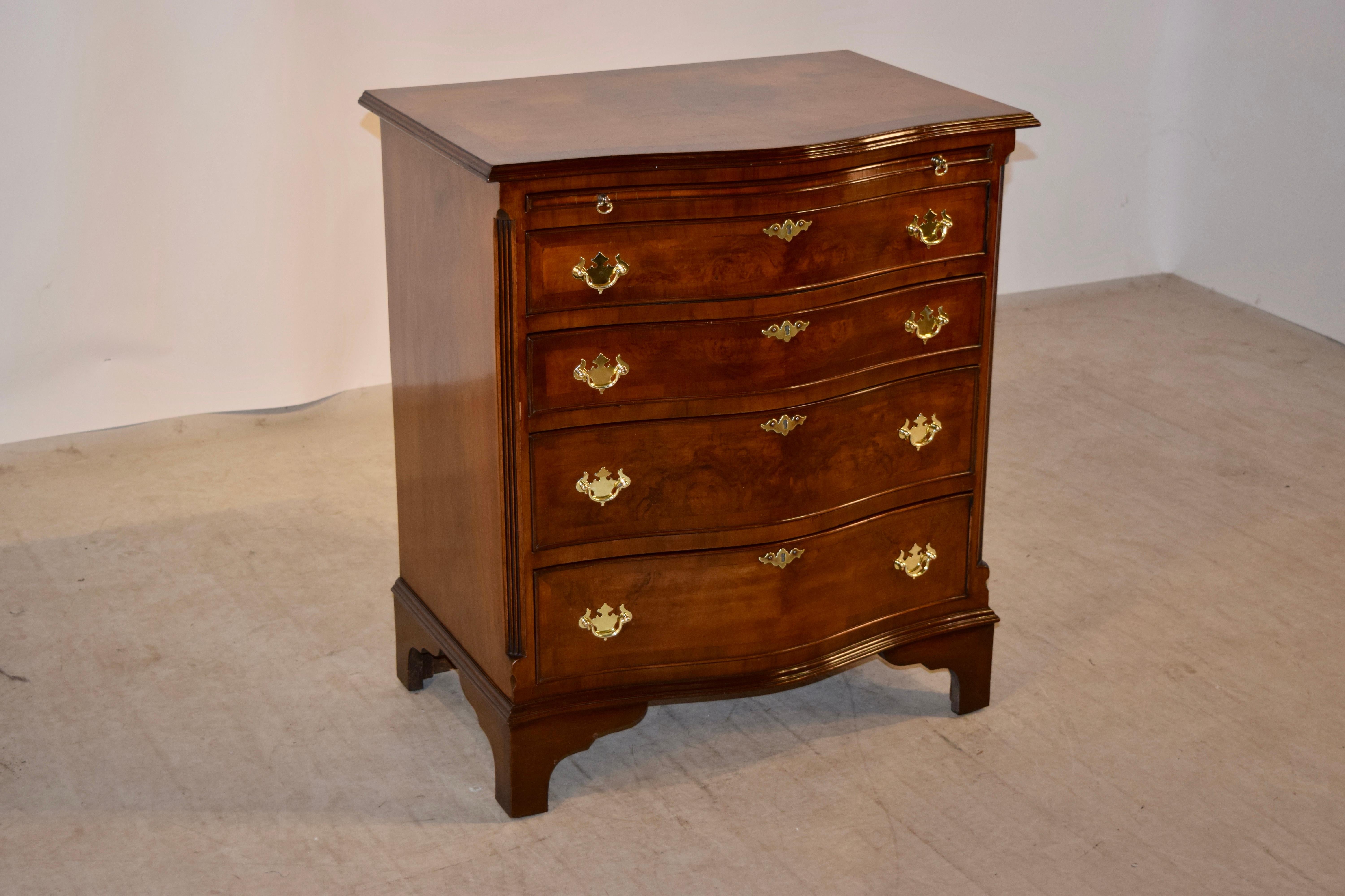 English serpentine side chest made from mahogany and walnut, circa 1920-1929. The top is banded in mahogany and has a central panel of burl walnut with a beveled edge, following down to a mahogany slide and four drawers, flanked by fluted quarter