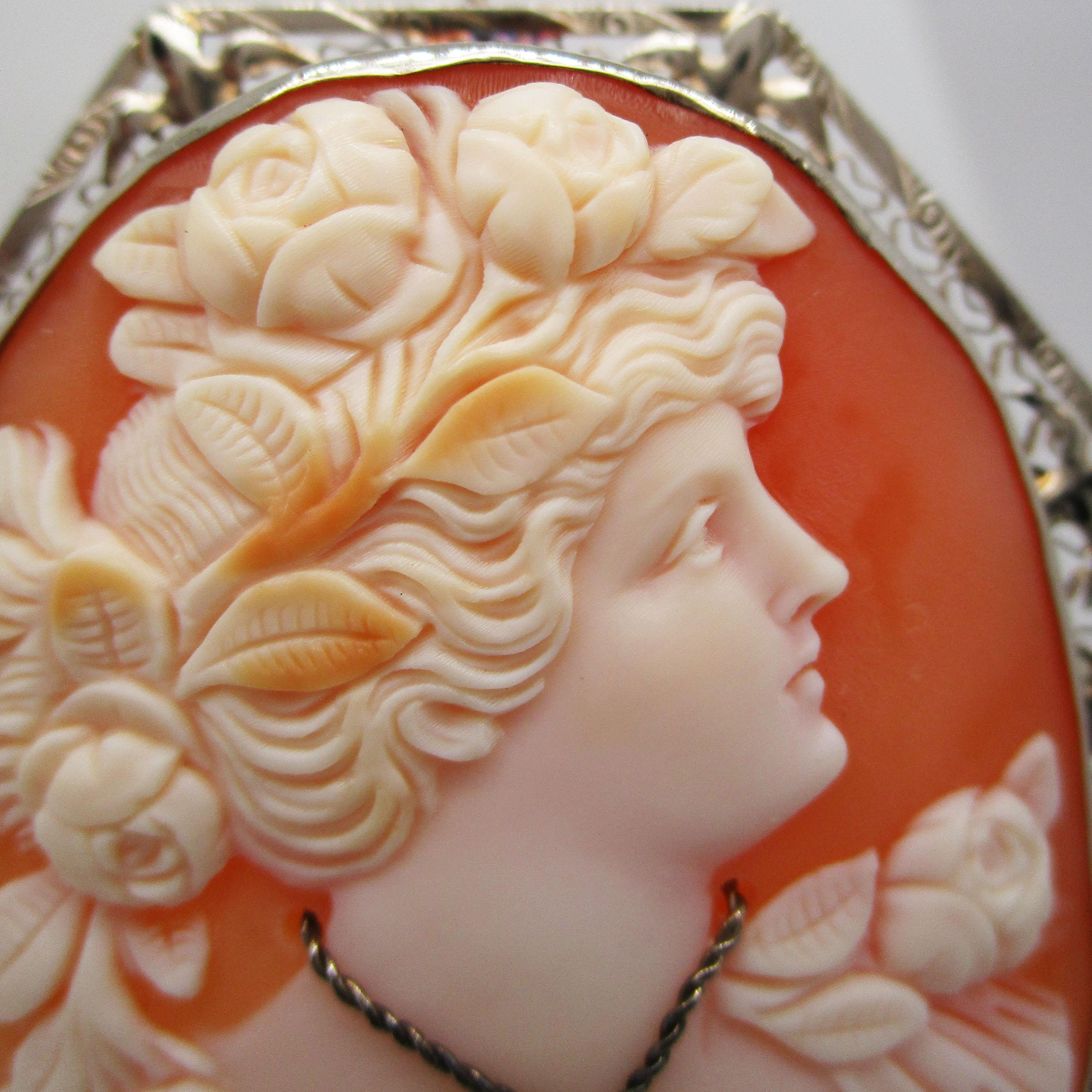 This is a breathtaking 1920s carved three-color shell cameo in a 14k white gold pin pendant. The cameo depicts a gorgeous left-facing woman with flowing hair and beautiful flower accessories. The woman is adorned with a delicate diamond necklace.