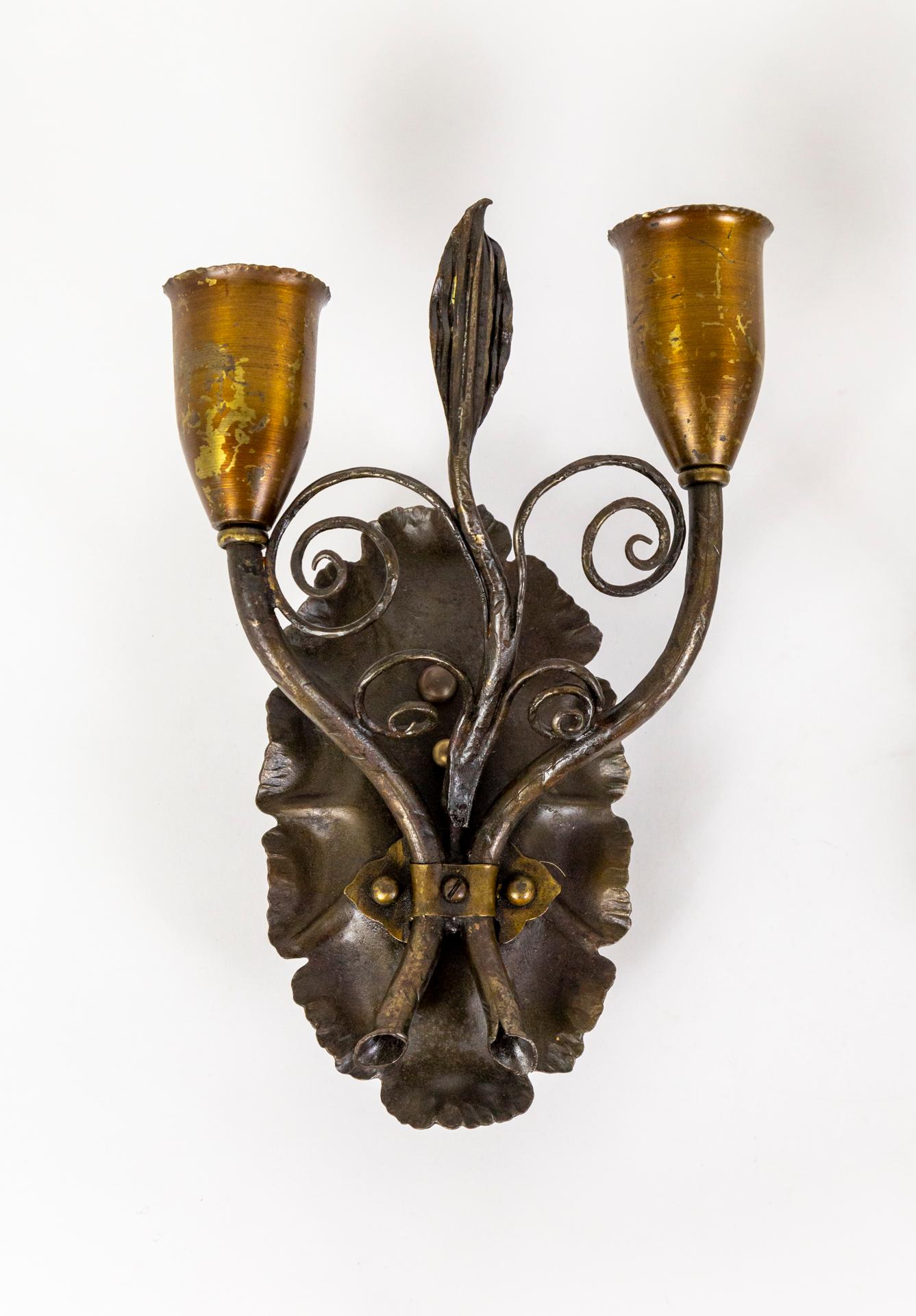 A pair of early 20th-century, iron and brass wall lights with two arms as elegant, trumpet stems fastened to scalloped oval back plates. They are detailed with a central, vine with a ribbed leaf and spiraling curls. They have a dark patinated body