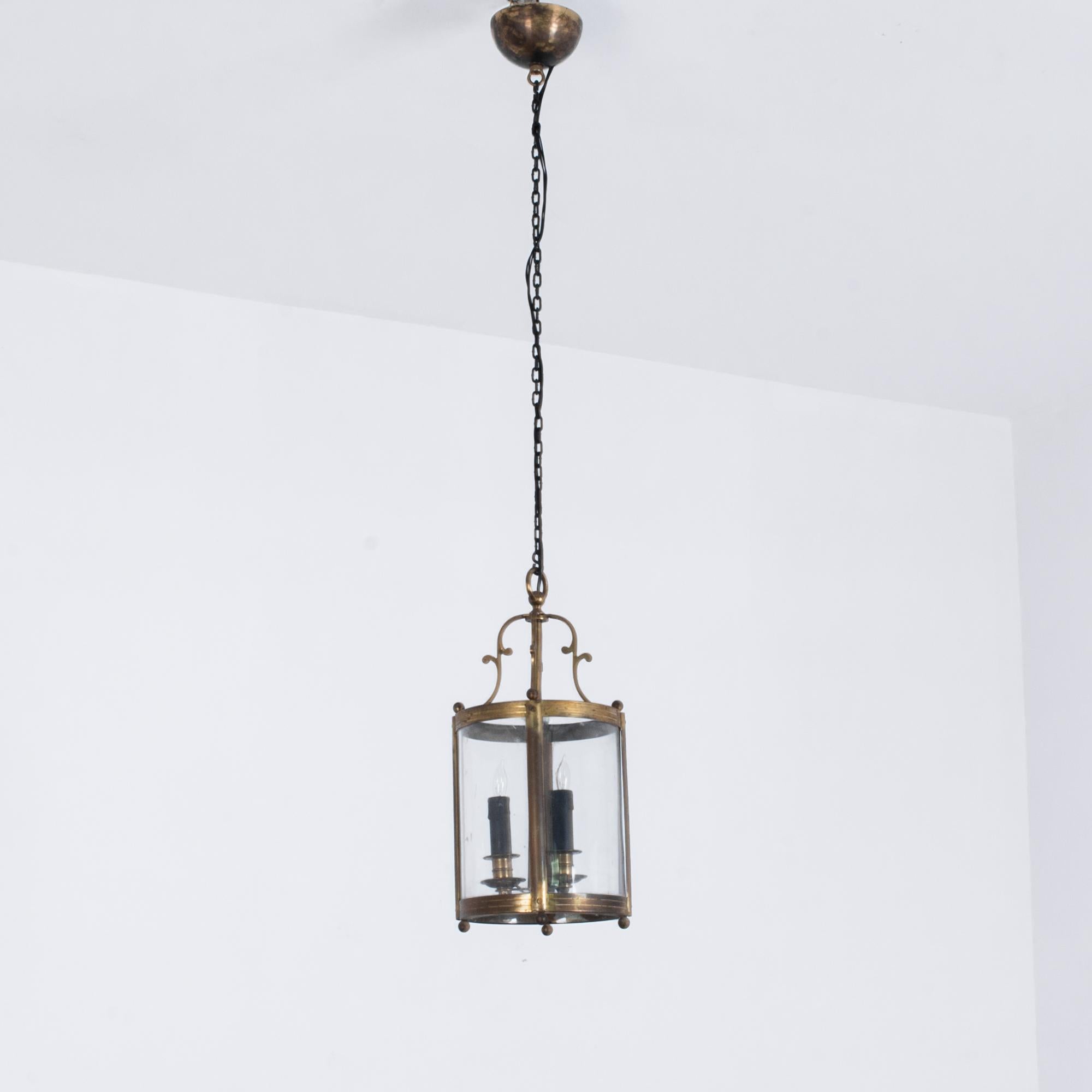 This brass lantern was made in France, circa 1920. It features a circular frame and two candle light bulbs, suspended from a ceiling rose. With the bell-shaped decorative elements above the lantern and small decorative spheres, the lamp will