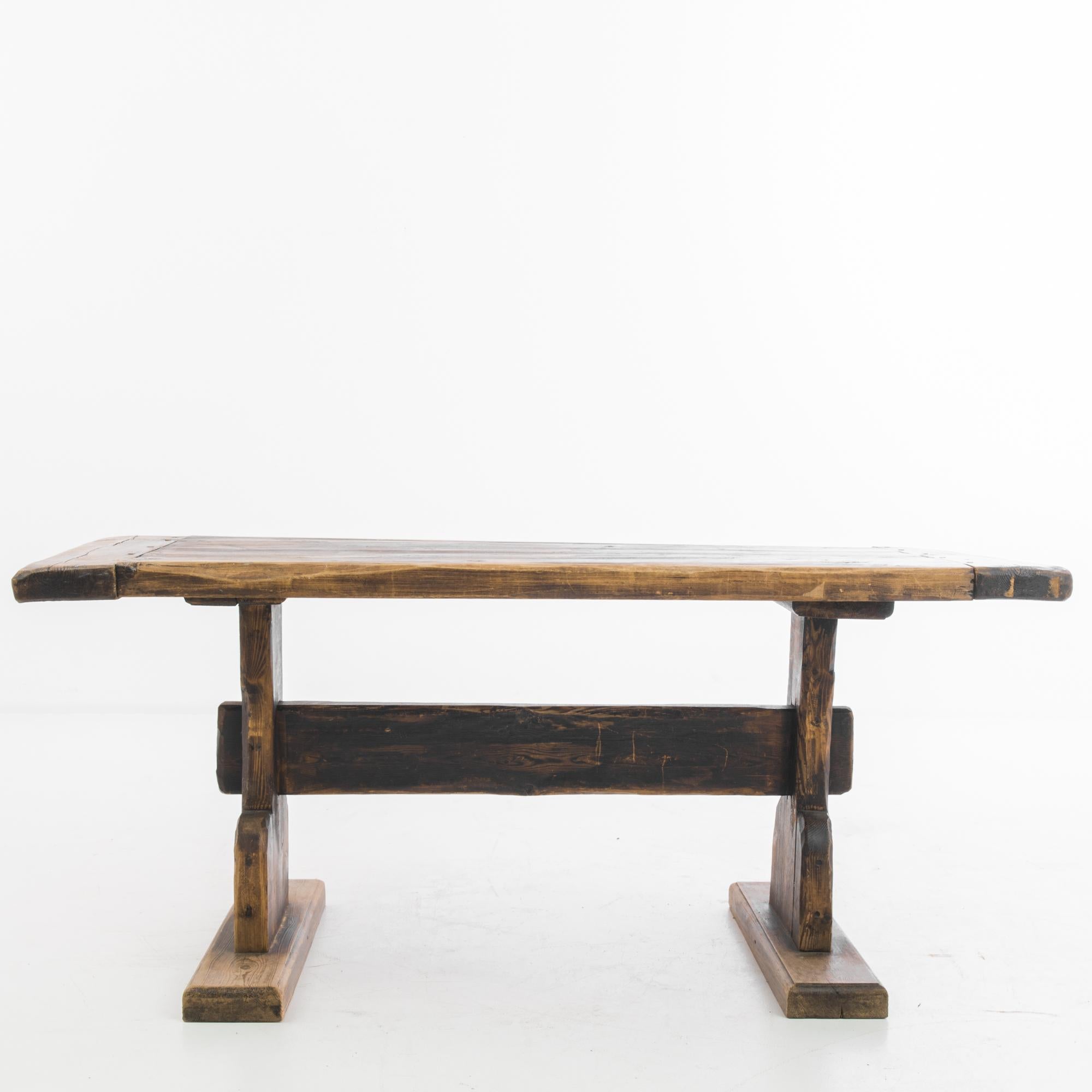 A wooden table from 1920s France. A rustic country silhouette with a dark, burnished finish. The broad slab of the tabletop rests upon two trestle legs, joined by a deep strut. The rough-hewn finish of the wood is mellowed by the warm color palette,