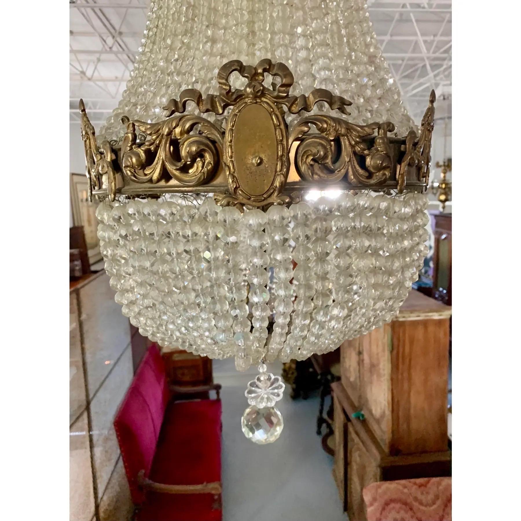 Araña francesa de cristal y latón de hacia 1920. Sartas de cuentas de cristal cuelgan de un dosel de latón coronado con un adorno calado. Debajo, un marco de latón está decorado con apliques de acanto con roleos y cartelas rematadas con cintas.