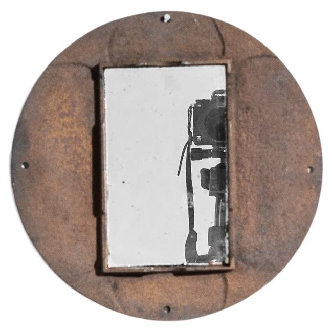 An iron mirror from 1920s France. An interplay of shapes and textures creates a weathered industrial aesthetic: a rectangular mirror is set inside a circular iron frame, oxidized to a dusky burgundy. The soft patina of the metal and the chipped
