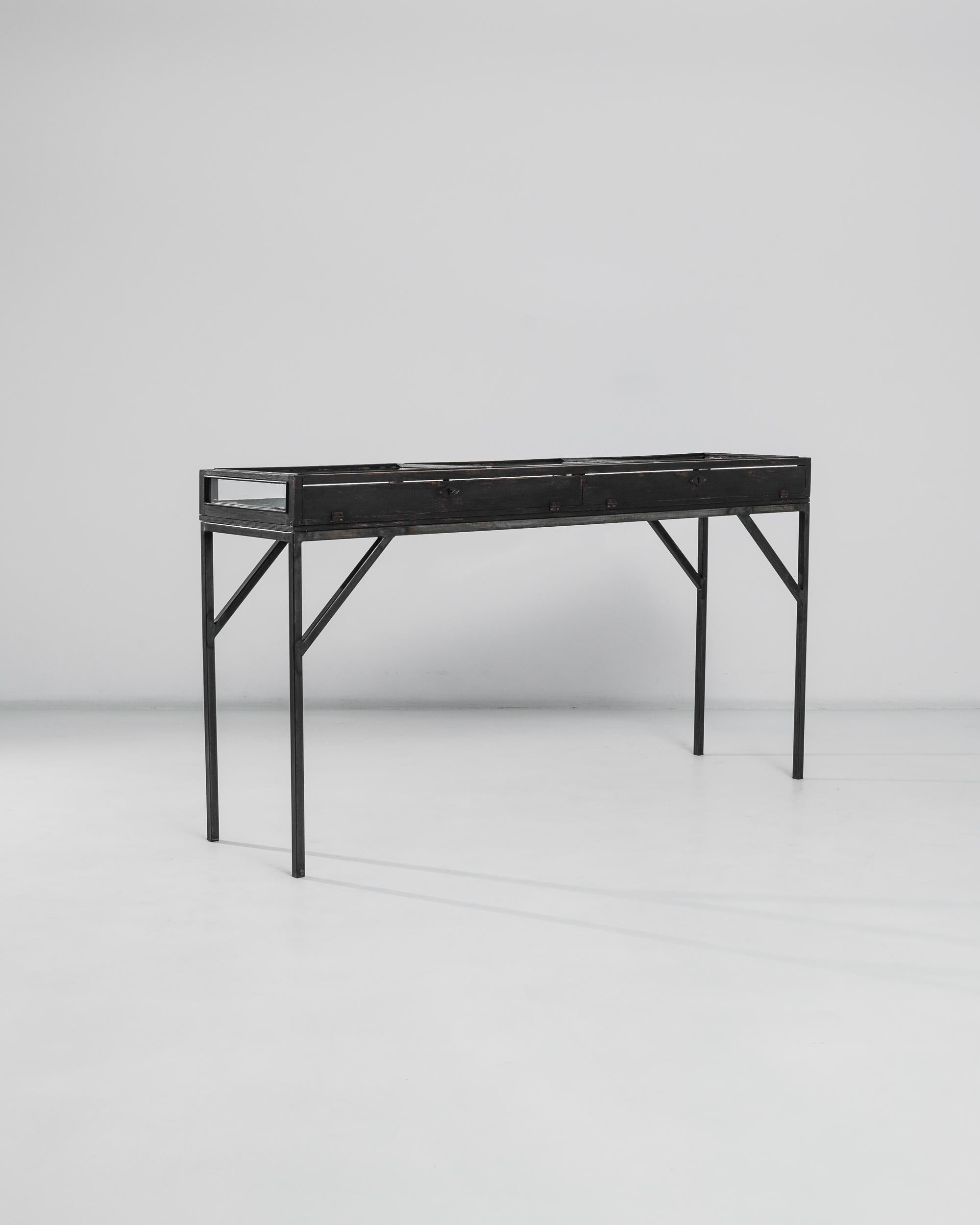 A metal and wooden display table made in 1920s France. Metal keys unlatch this minimal vitrine, allowing access to its delicate interior. A bright brownish red wood hue is revealed in flashes upon the painted exterior of the table, producing a