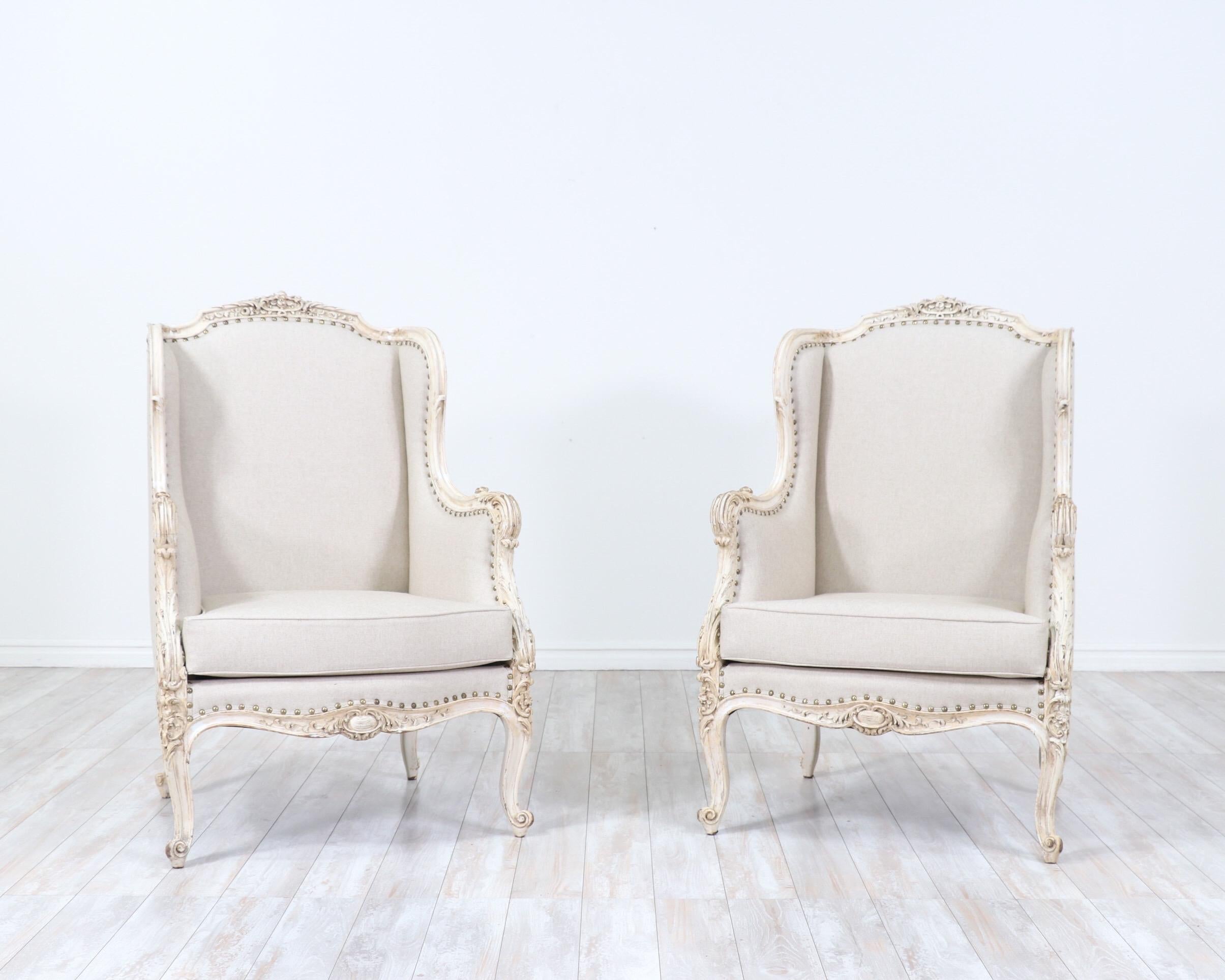 Beautiful, French 1920s pair of bergeres in the Louis XV style.

The chairs feature intricately carved wooden frames in a creamy white distressed paint finish, new canvas linen upholstery in a tan-oatmeal color, loose seat cushions and antiqued