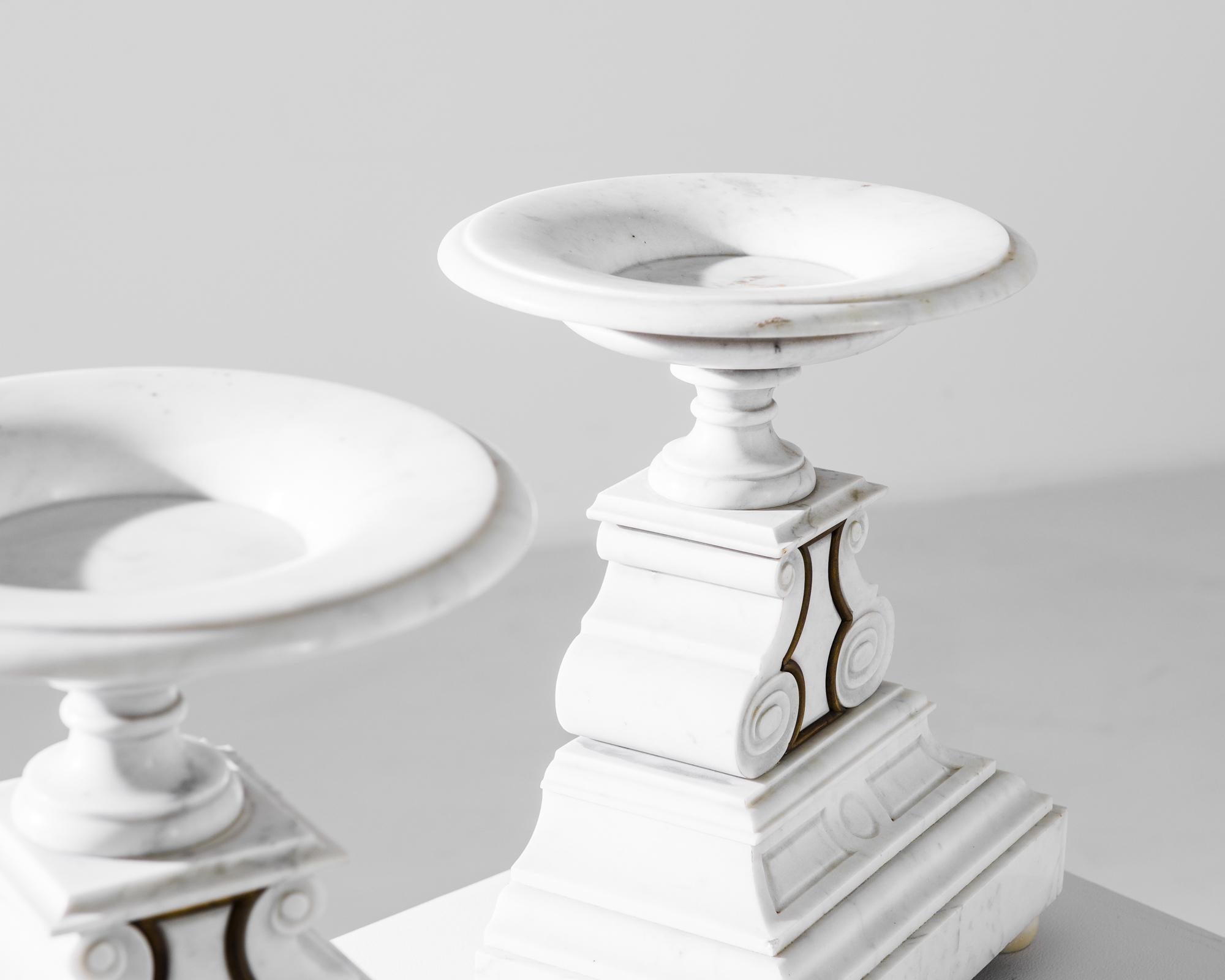 An elegant pair of marble pedestals from 1920s France. A tapered pyramidal base in pristine white marble is crowned by a circular dish. Carved scrolls, accentuated by gilded metal accents, add a touch of Beaux Arts embellishment — combining the