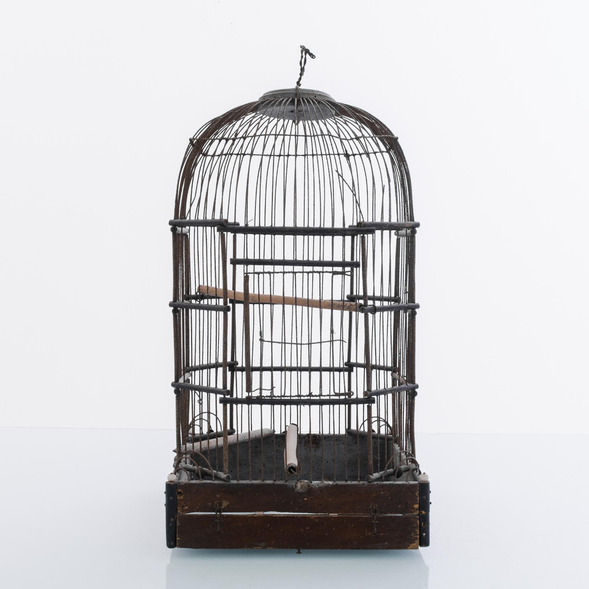 This metal birdcage with a square wooden base was made in France, circa 1920. Corrugated and smooth rods alongside thin wires were used to construct the cage, which features a dome top. Light brown wooden perches are fitted in the center and at the