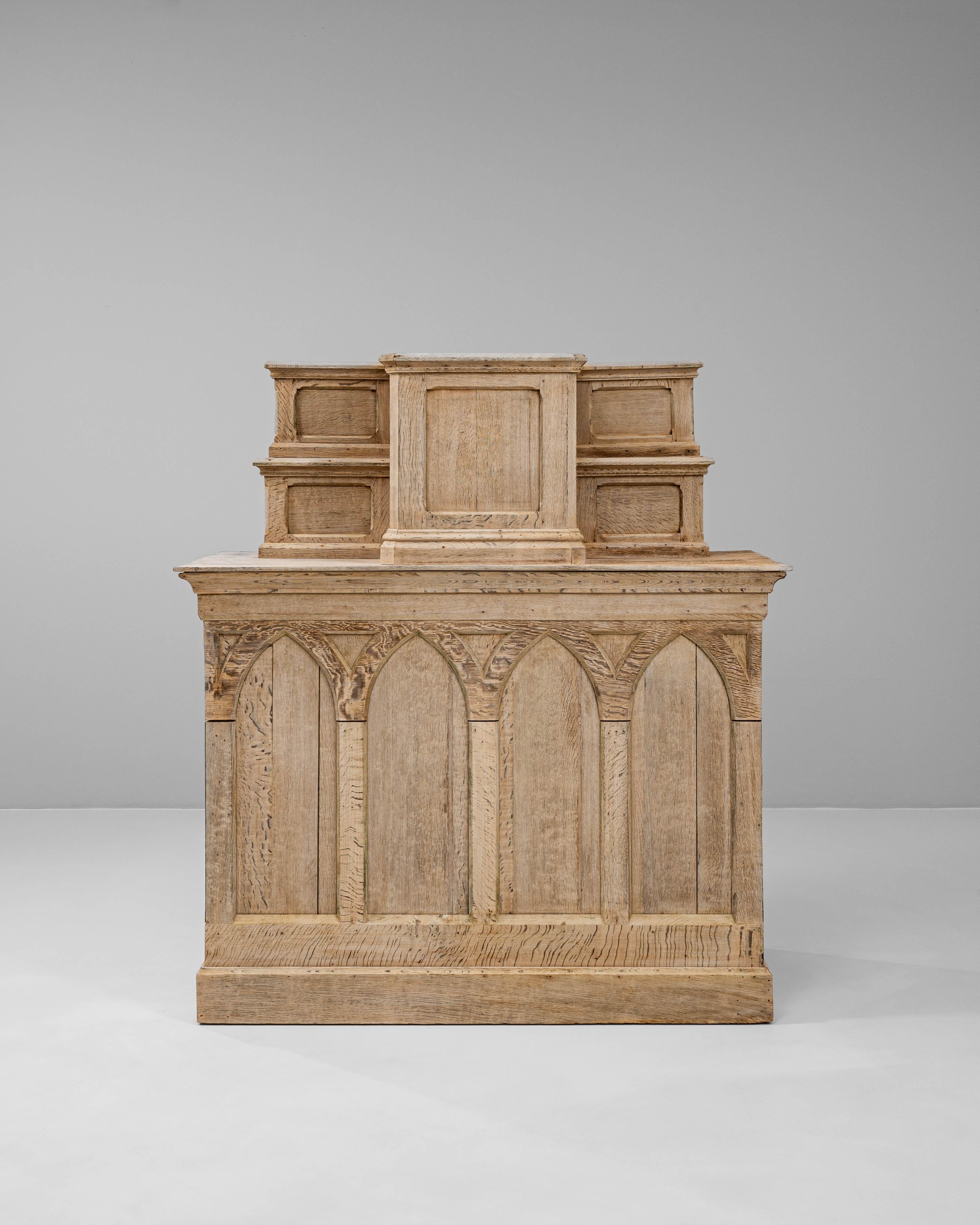This 1920s French oak display cabinet is a striking example of Gothic Revival craftsmanship, distinguished by its pointed arch paneling and stepped upper tier. Carved from solid oak, the cabinet features a pale, natural finish that highlights the