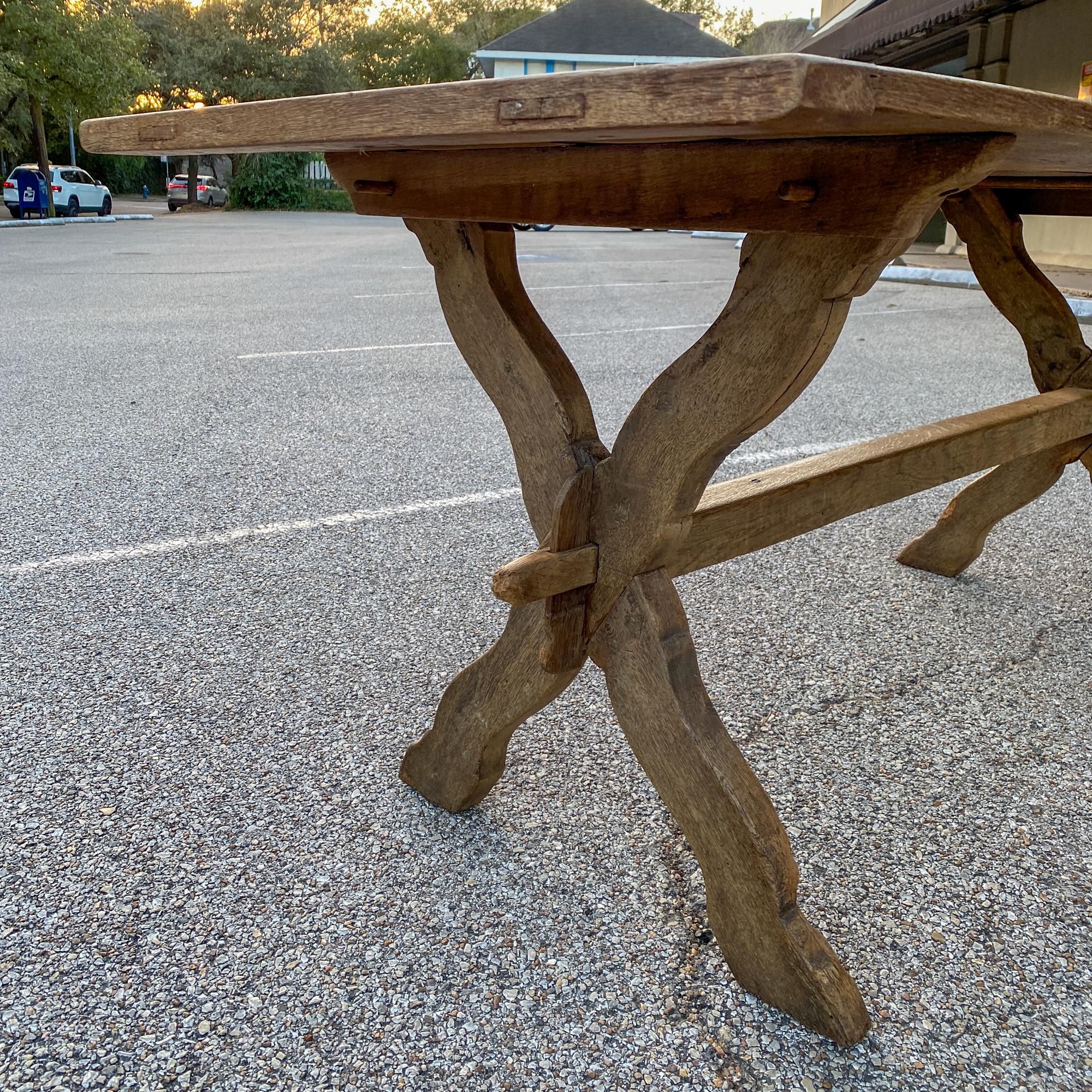 This 1920s French oak trestle-style farm table features an X-shaped base with a center stretcher. This piece is crafted with wooden pegs that hold everything together, allowing it to be disassembled completely and packed flat, making this an ideal