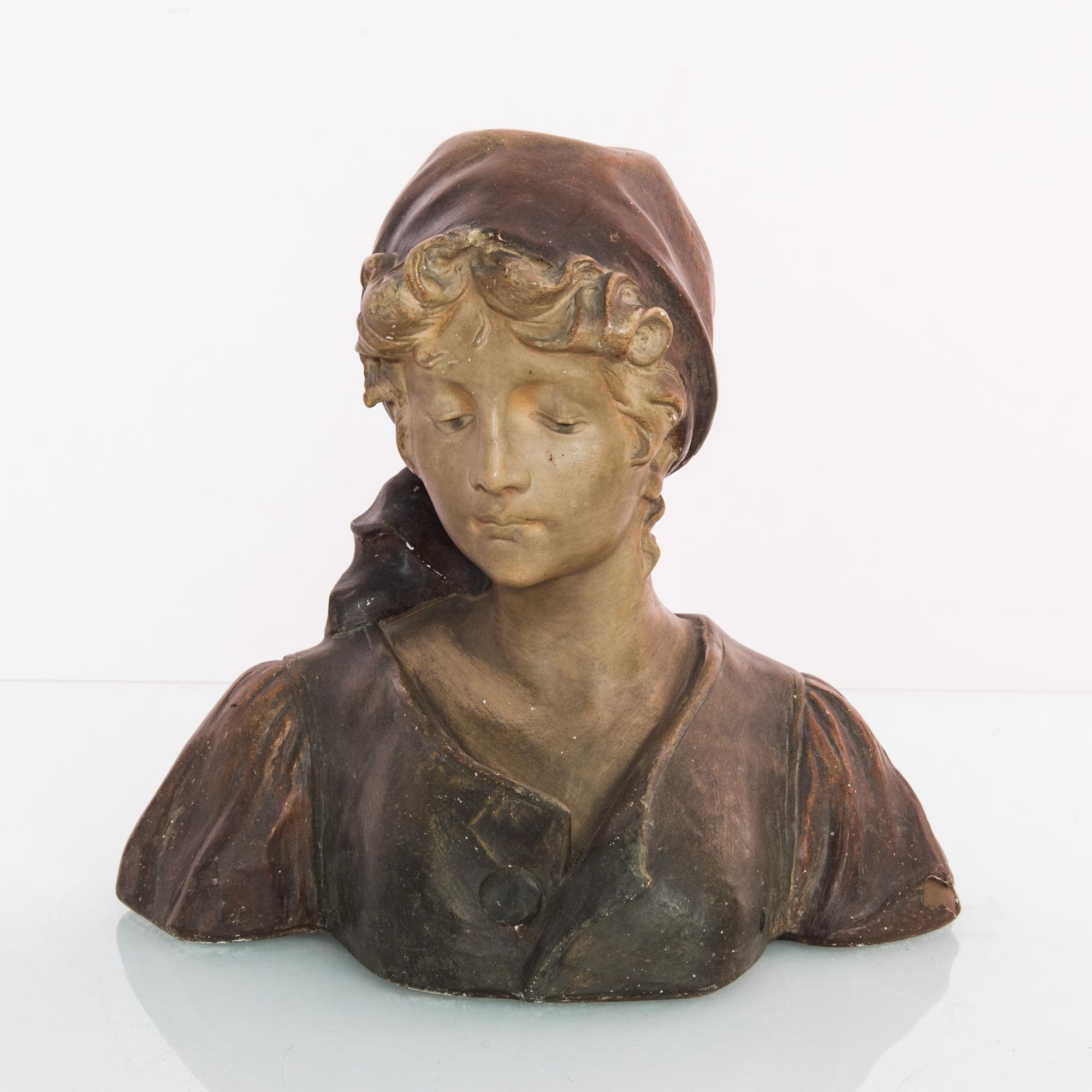 This ceramic bust was made in France, circa 1920. The refined sculpture shows a young woman, with a scarf covering her hair. A dour pose shows her looking down, in contemplation, with a beautiful beige and dark brown patina patina.