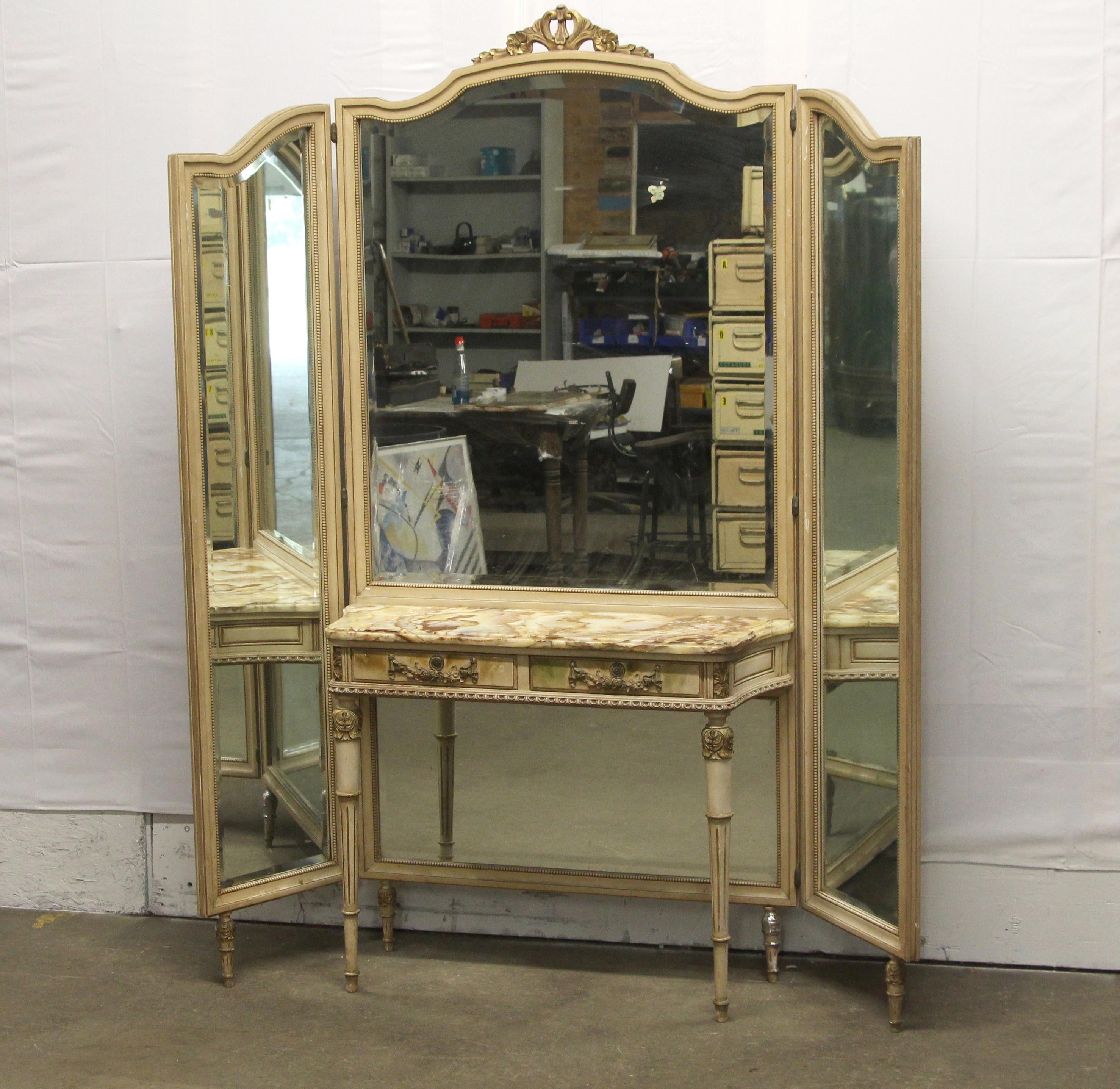 1920s French Provincial carved and hand painted wood vanity table with two drawers and an attached tri-fold mirror. It features a delicate onyx top table with orange, yellow and white veining. There is damage to the one leg. The dimensions are Table