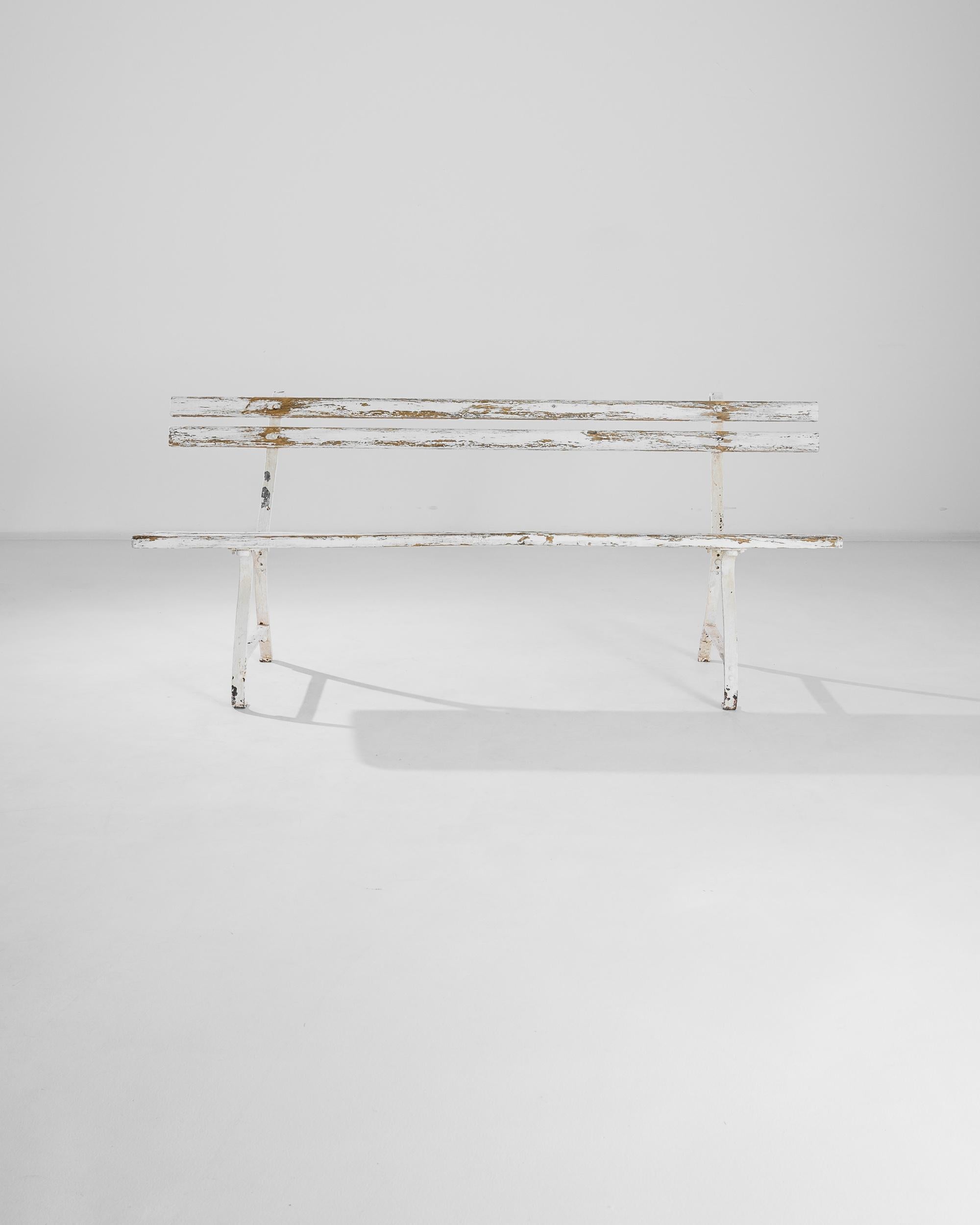 This antique wooden bench with metal frame was produced in France, circa 1920. Nicely distressed, the charming bench offers a slender silhouette, invigorated by splay legs curved inward. The white paint flaunts a mature patina, revealing the