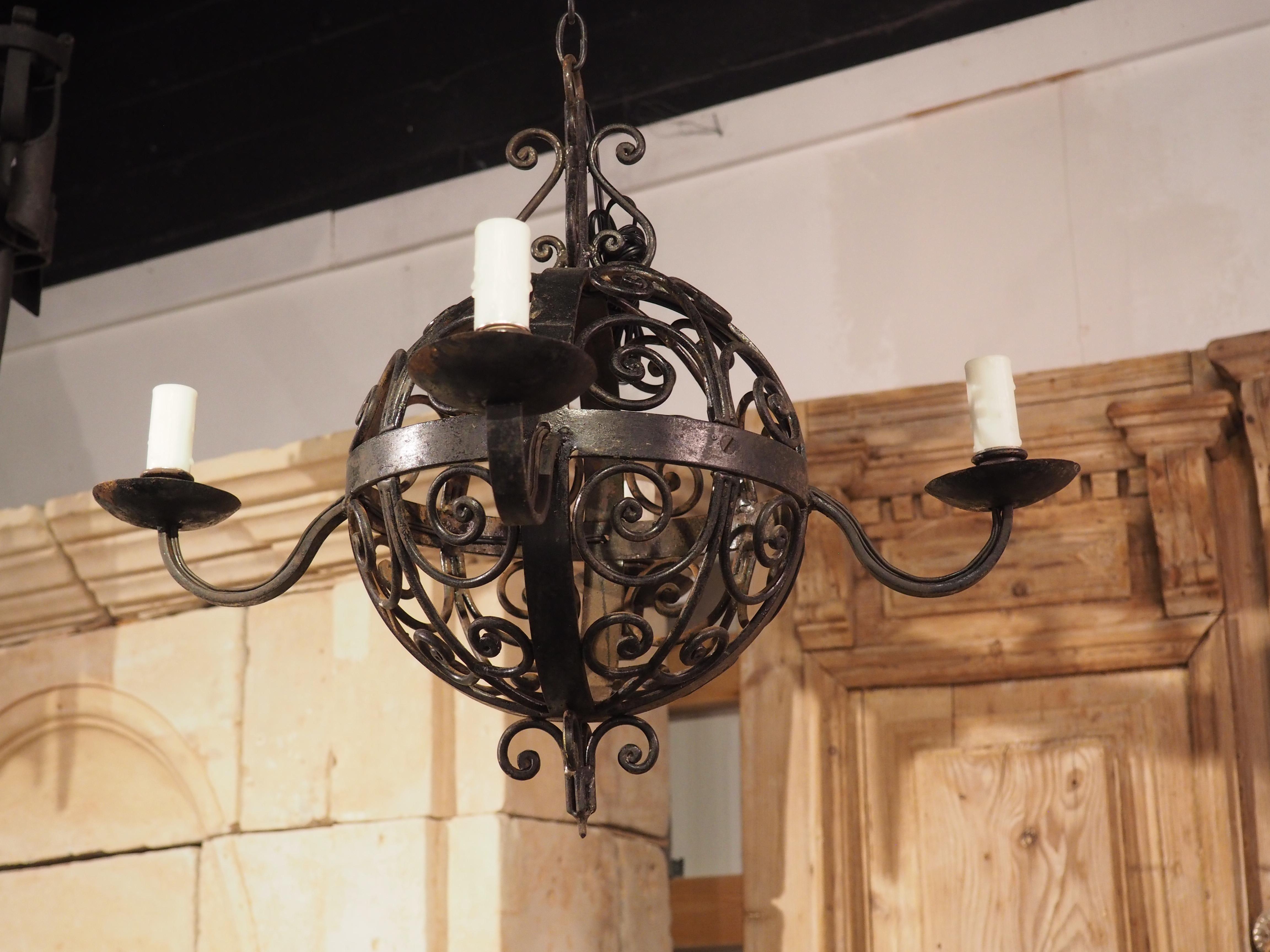 Crafted in France during the 1920’s, this wrought iron ball chandelier features four saucer sconces on top of S-scroll arms. Each of the arms is affixed to a band that circumnavigates the open sphere laden with scrollwork. The highly worked sphere