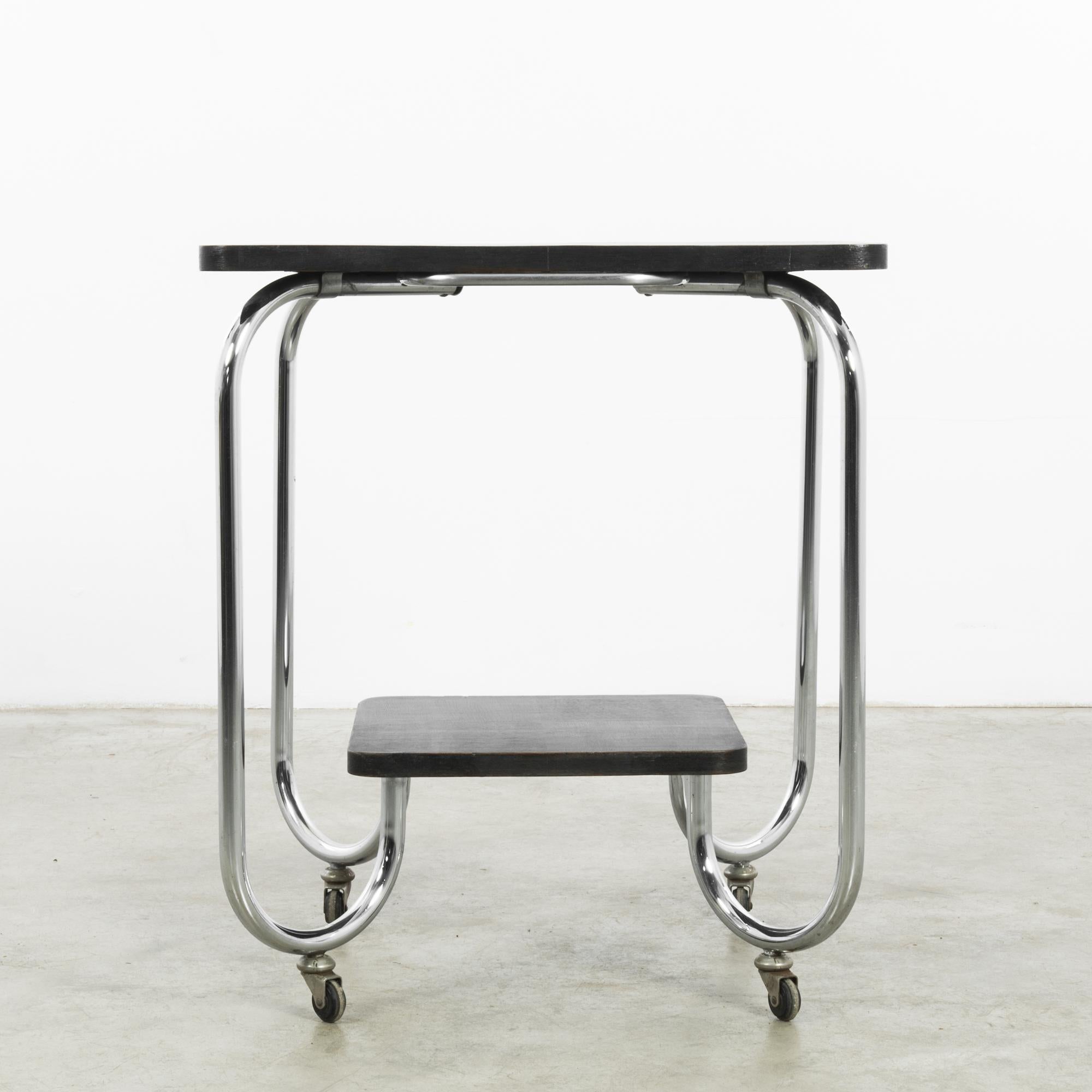 This 1920s German Metal Table boasts a classic vintage design, featuring a sleek, tubular metal frame that supports a rich wooden top. Its compact structure is heightened by the functional addition of wheels, making it a versatile piece for any