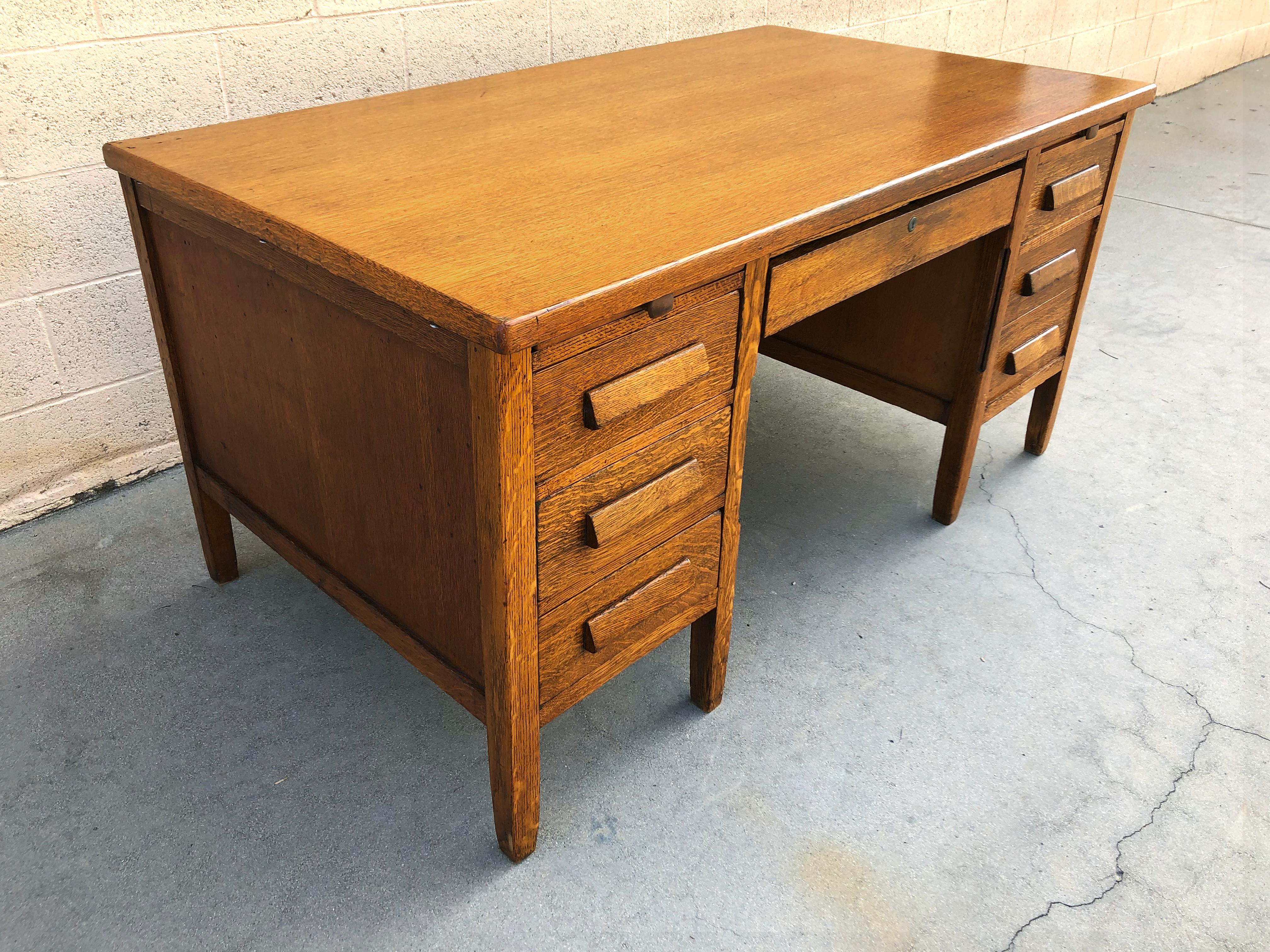 1920s teachers desk made of solid golden oak. Well made in a straight and simple Arts Crafts/ Mission style. Features double pedestal configuration with four storage drawers, a filing drawer, center utility drawer and two pull-out writing trays.