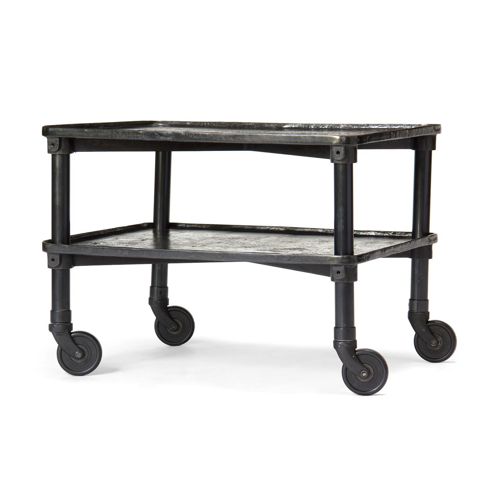 A patinated cast iron two-tier industrial table on single wheel casters.