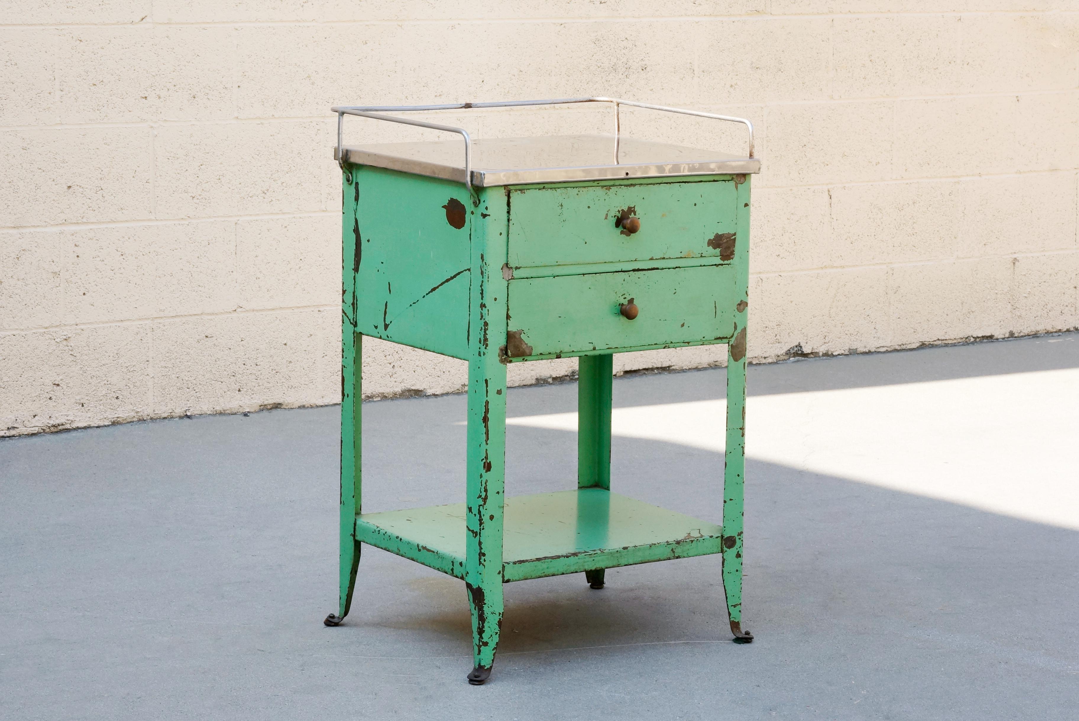 1920s industrial side table with stainless steel tray top. Possibly from a hospital setting. Features two drawers and bottom shelf. Weathered original green patina and wood pulls. 

Dimensions: 21