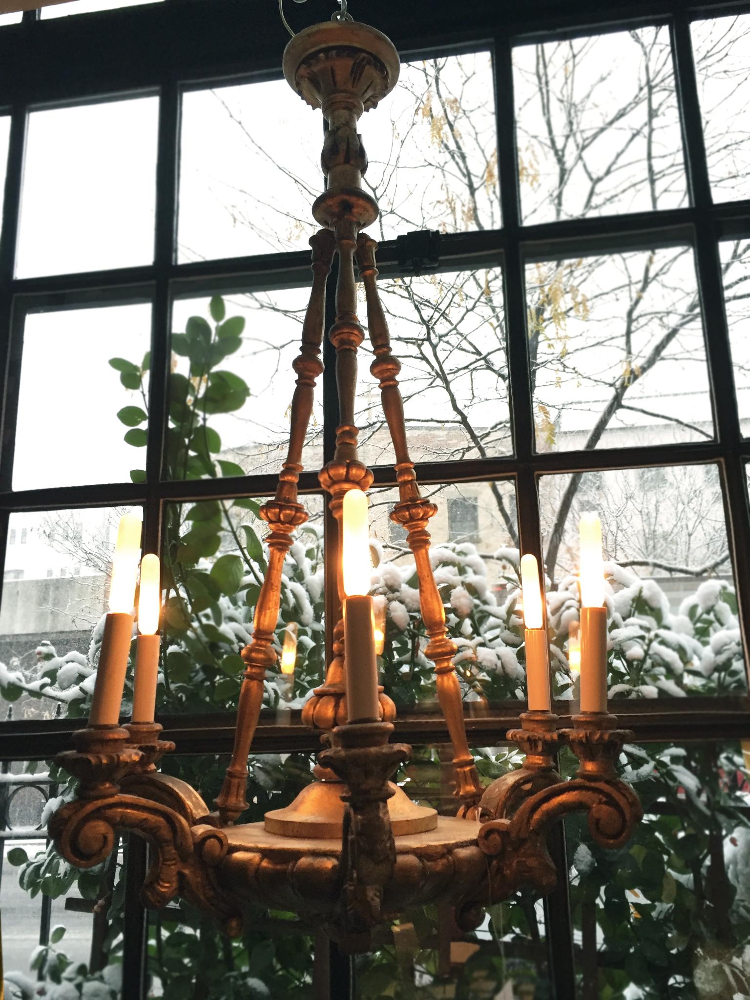 This 1920s Italian chandelier is comprised of wood with a gilt finish. The piece has a Baroque sensibility with its dramatic scrolled arms, bobeches, spindle frame, and canopy. The spindle frame leading up to the canopy is collapsible and hangs