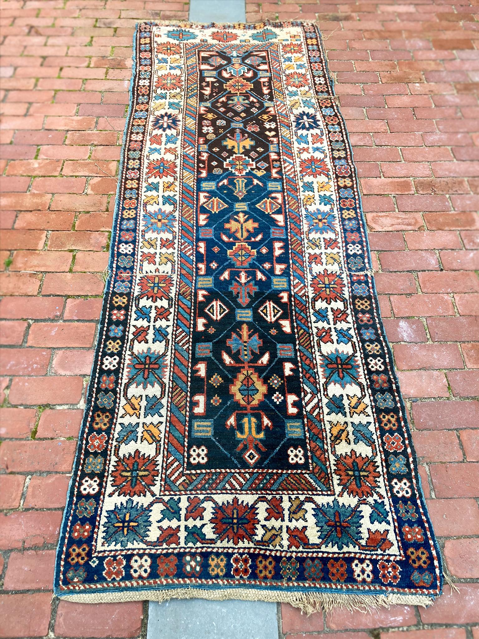This 1920s Kazak runner rug has a vibrant palette that consists of red, ocher yellow, navy and light blues, and ivory. These colors are arranged intricately in stylized floral and geometric patterns. The rug has been professionally cleaned.