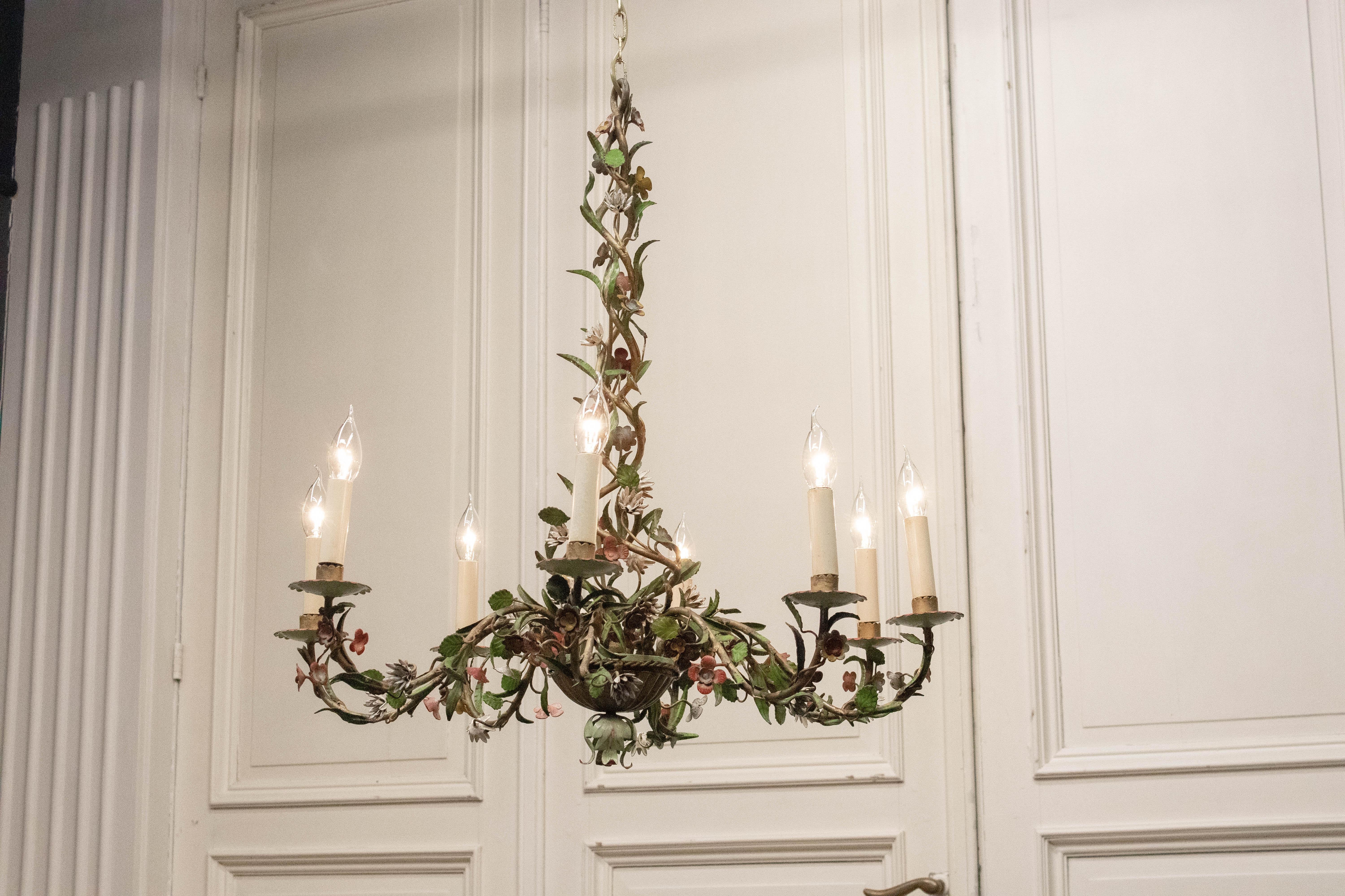 Large 1920s French tole chandelier featuring an ornate, hand-crafted composition of painted metal flowers, leaves, and winding vines. This impressive piece showcases six scrolling arms, each supporting a candle-style light, and a richly detailed