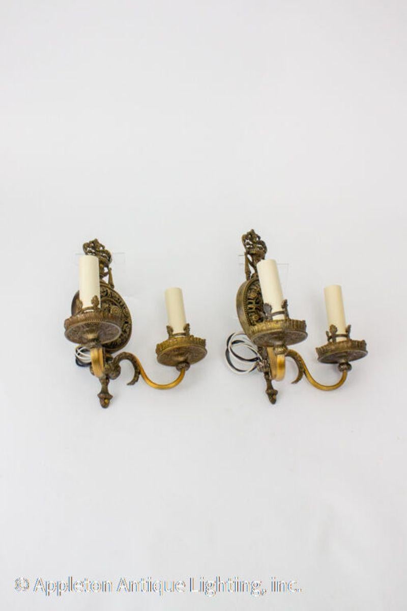 A pair of 1920’s Lion Electric vintage sconces. Two arm sconces with ornate gold colored metalwork over a subtle dark green backplate. Design is of an urn with floral arrangements, organic elements and draping. Original candle cover rings, unique to