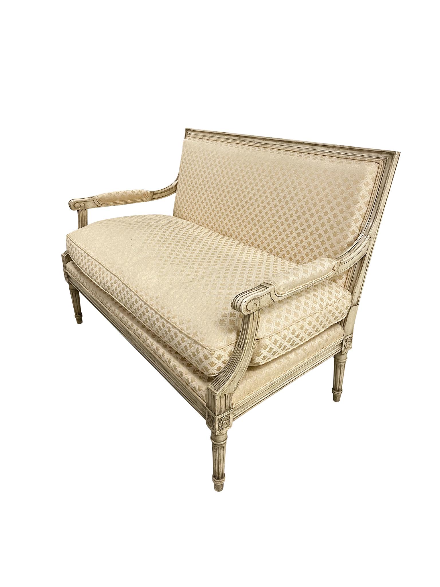 This classic Louis XVI-style settee was made in the 1920s. Its comprised of a painted, wood frame upholstered with fleur-de-lis patterned fabric. The arms are padded with the same fabric and meet the legs in a curved, fluted
