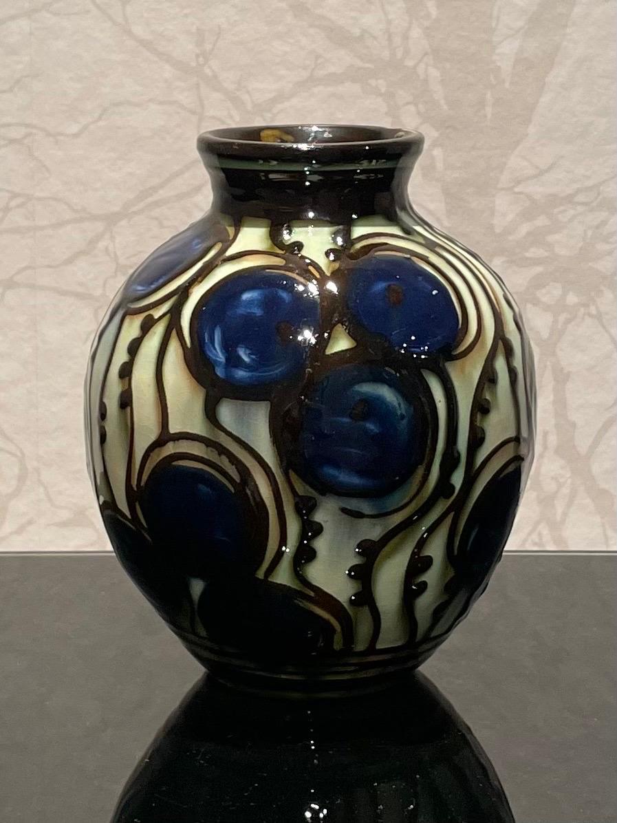 This is the 1920s Danish 18 cm high ceramic vase by Herman Kähler. 

It comes with a globular body. It has a high glossy surface, a beautiful cow horn glazed pattern with thin climbing plants and royal blue flowers on a beige background. 
The gently