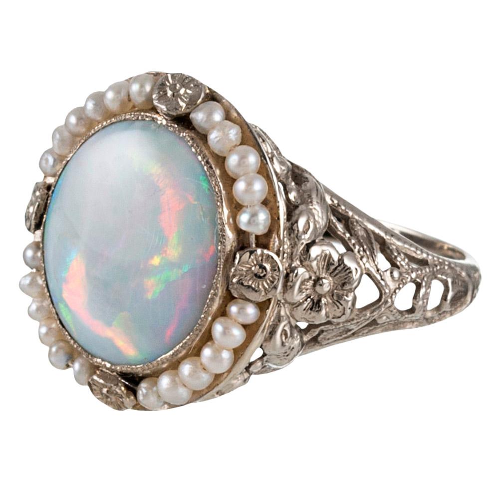 Feminine and delicate, this 14 karat white gold filigree ring is set with a lustrous opal cabochon and framed with seed pearls. Note the flower accents at the compass points and the gently textured bezel. The low profile of the design will allow