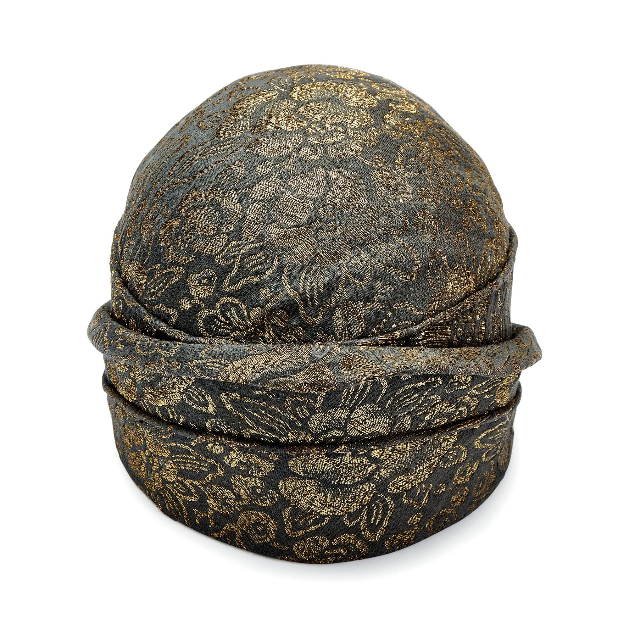 This highly collectable hat was designed in the 1920s and is tailored to a classic cloche shape. An expensive and well made couture piece, it is crafted from a charcoal lamé detailed with gold metallic thread. This shimmering thread forms floral